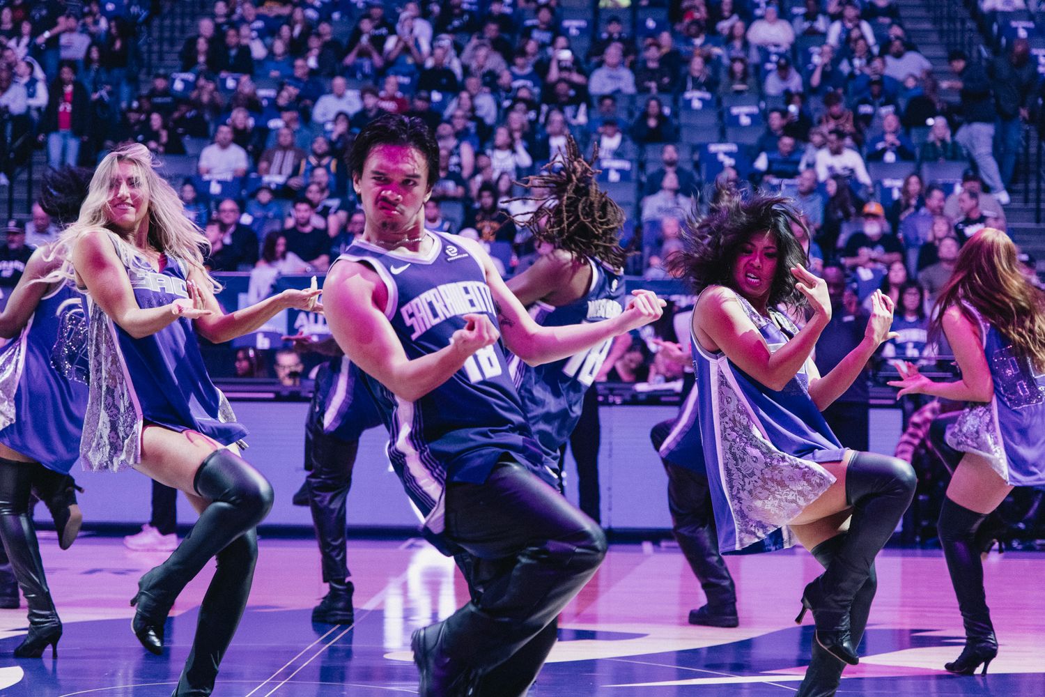 NBA dancers perform energetic routine on basketball court in purple uniforms before packed arena crowd.