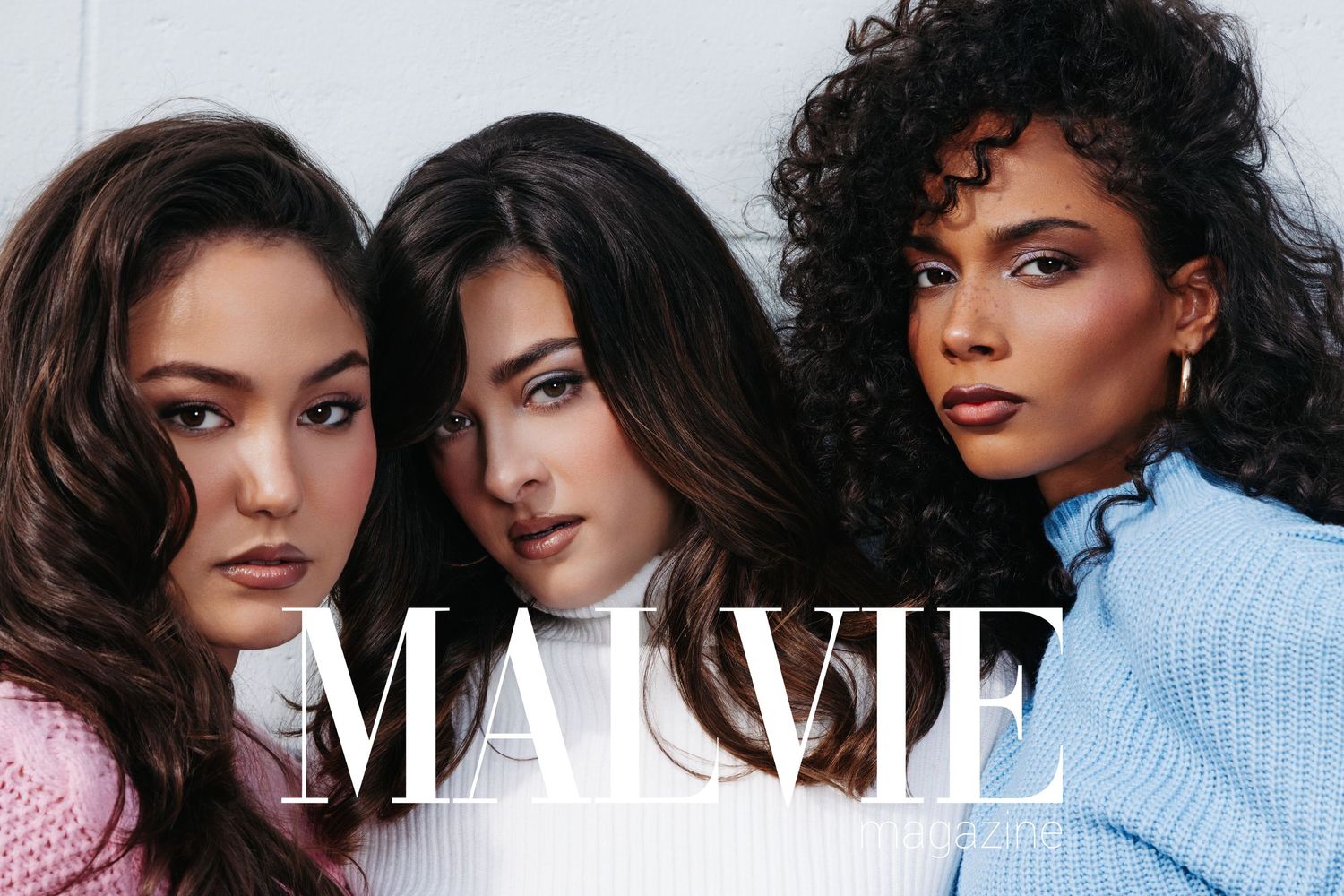 Three diverse women models pose together for Mauvie Magazine, wearing pastel sweaters in pink, white, and blue.