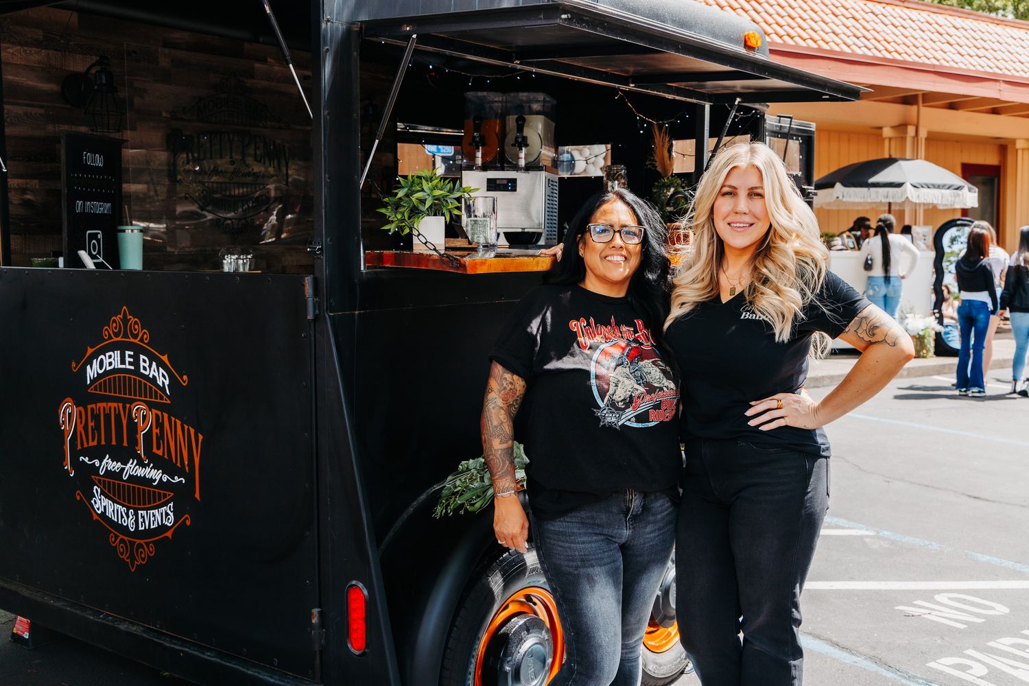 Two women in black shirts posing in front of the Rusty Pony food truck at an outdoor event.