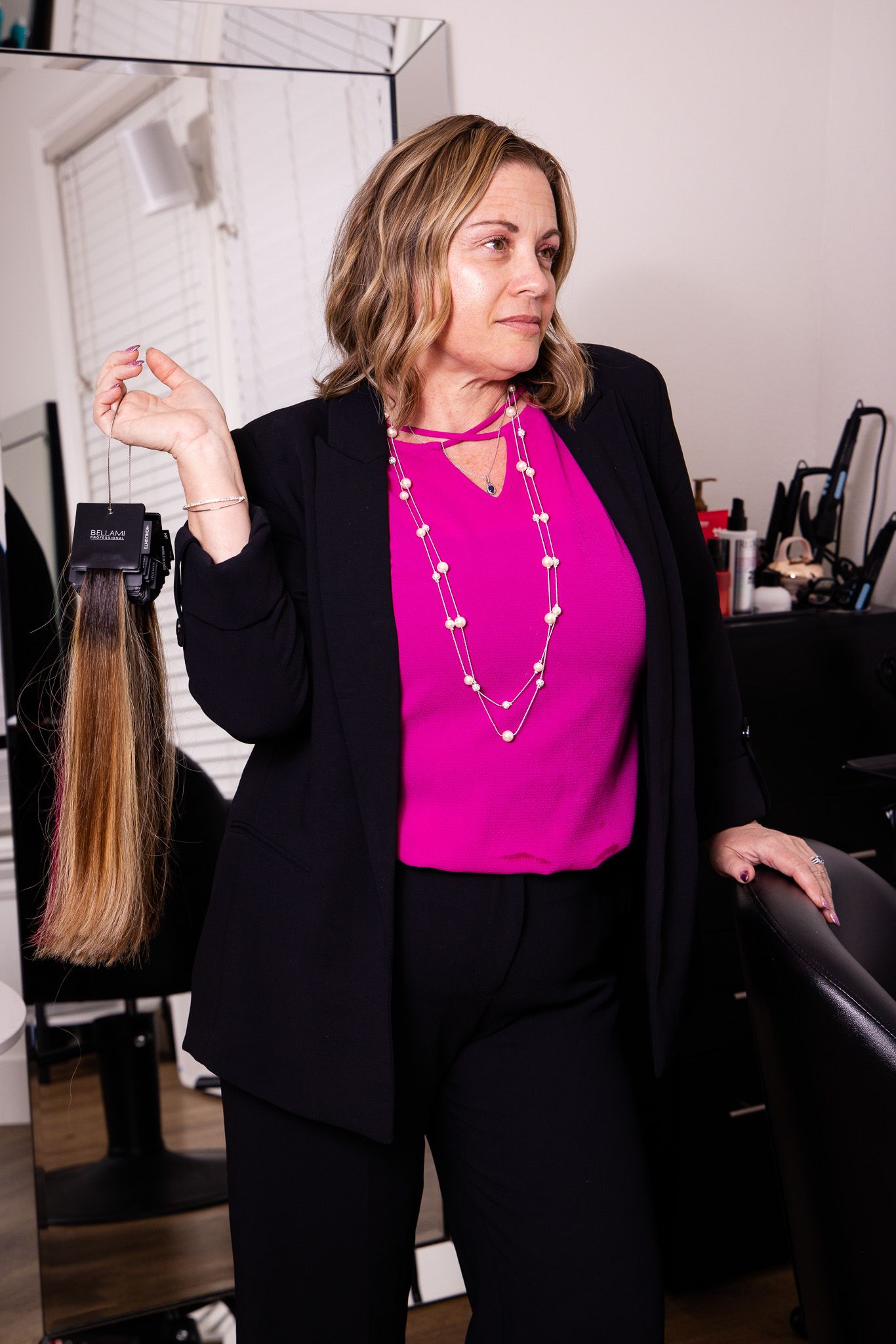 Stylist in black blazer and magenta top demonstrates hair extension while working in salon setting.