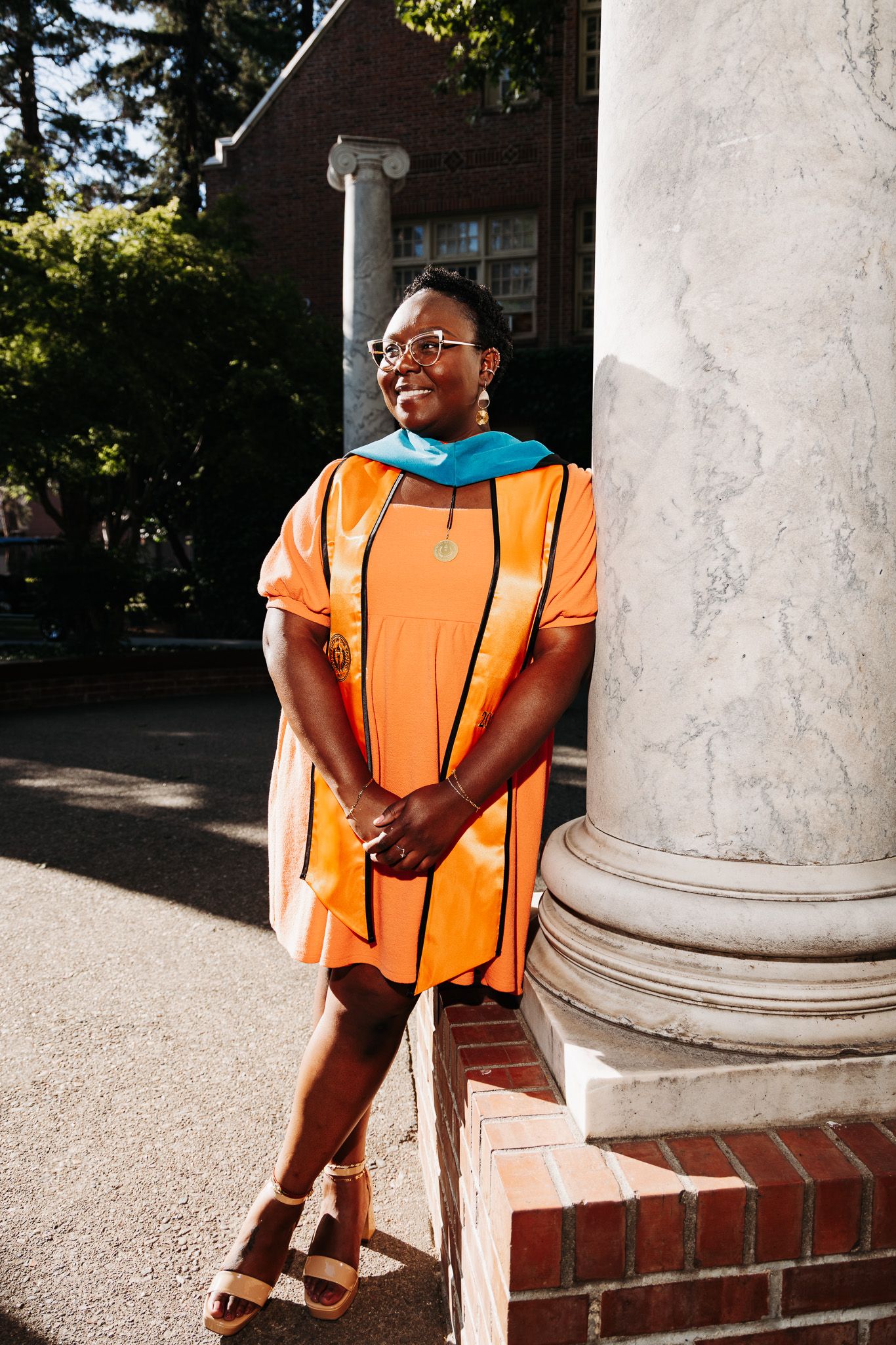 A person wearing a bright orange dress sits on white stone columns with brick steps in an outdoor setting.