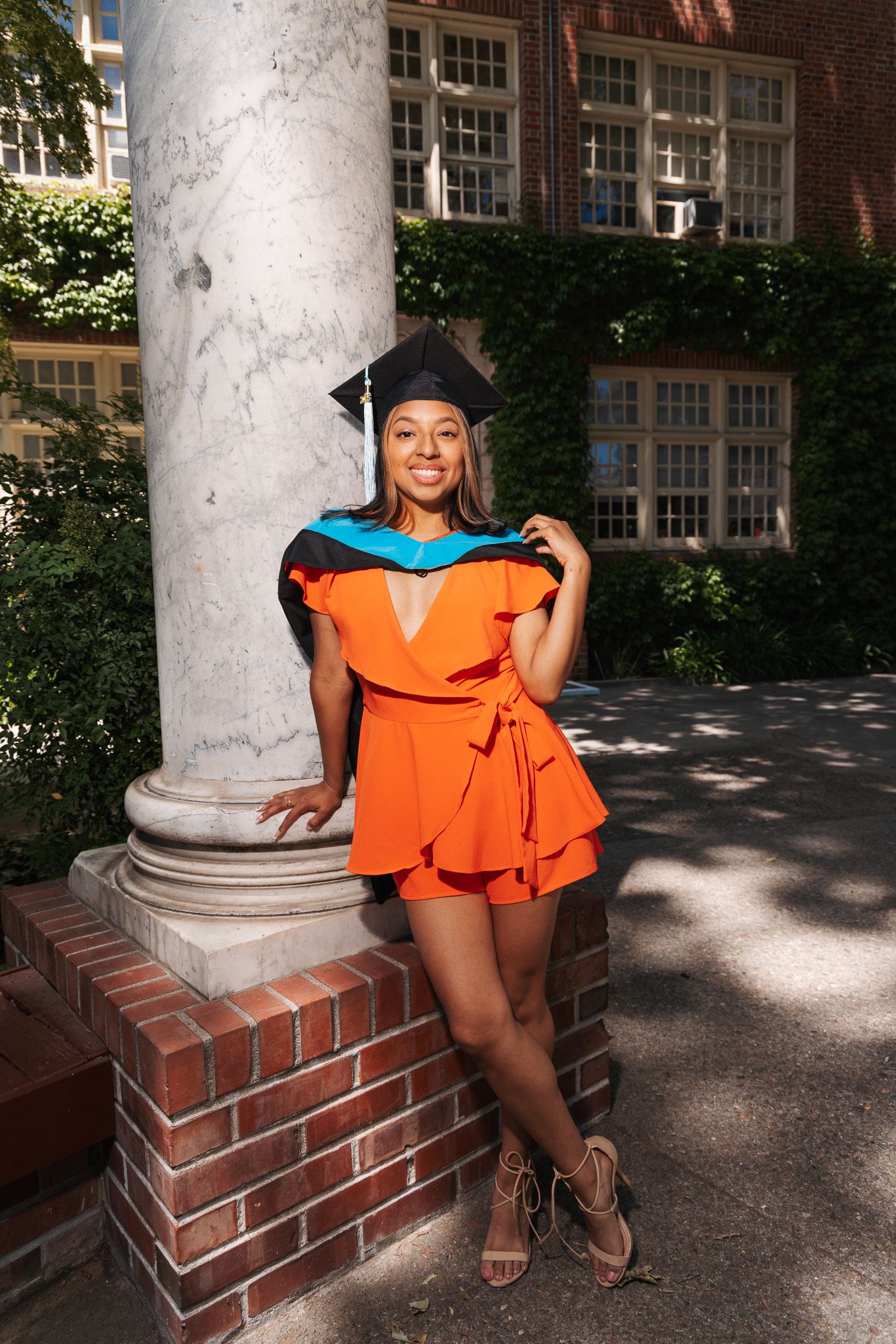 Graduate in orange ruffle dress poses beside white stone column for celebration photoshoot on university campus.