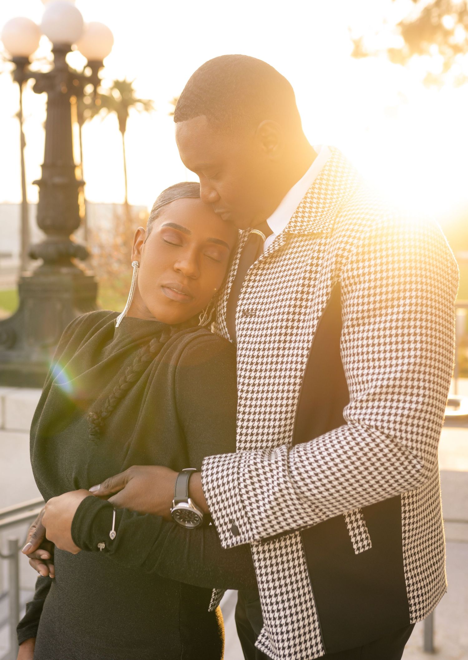 A couple embraces in warm sunset light while wearing black and checkered outfits against an urban backdrop.