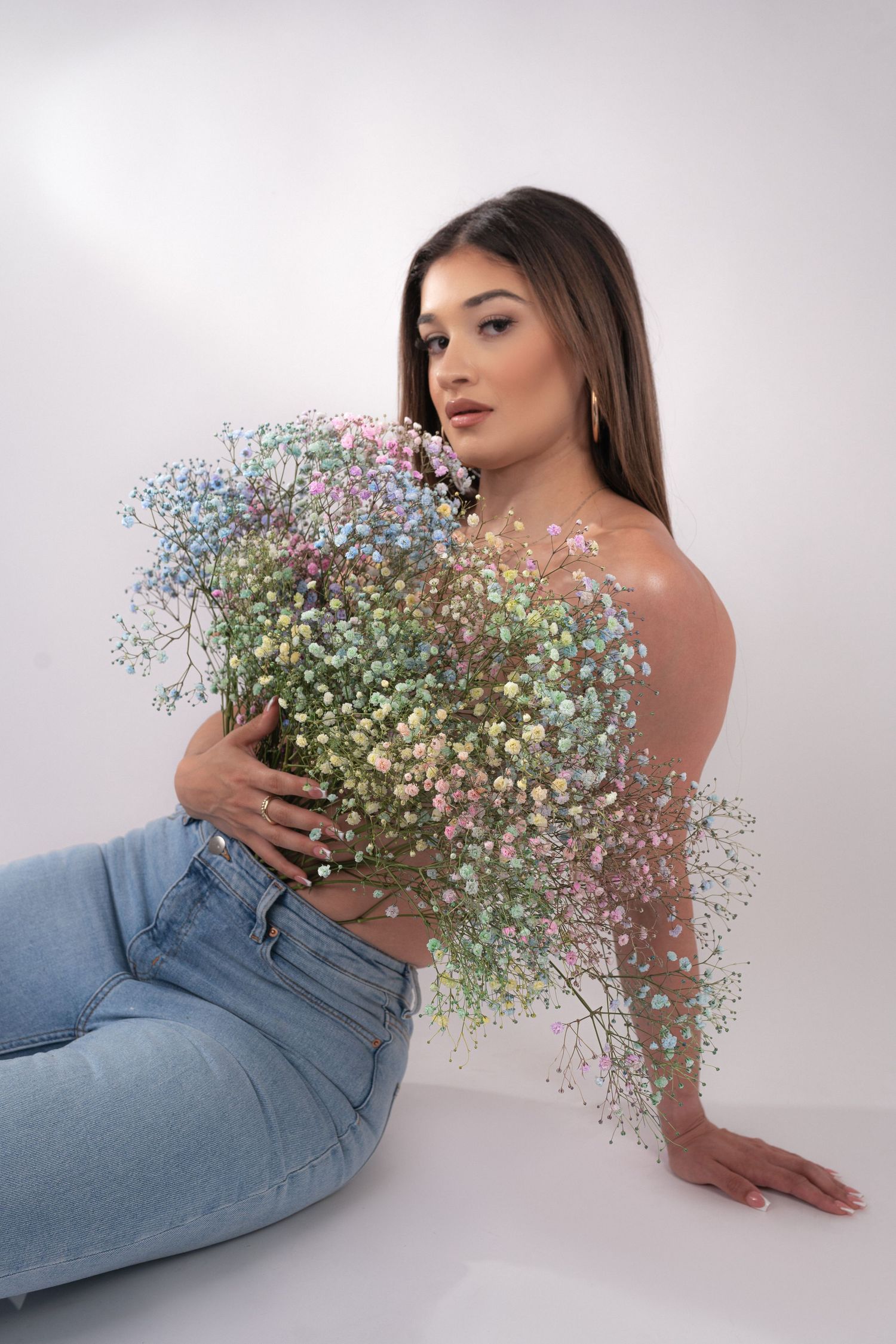A woman in light blue jeans lies on a white floor holding delicate baby's breath flowers in an artistic pose.