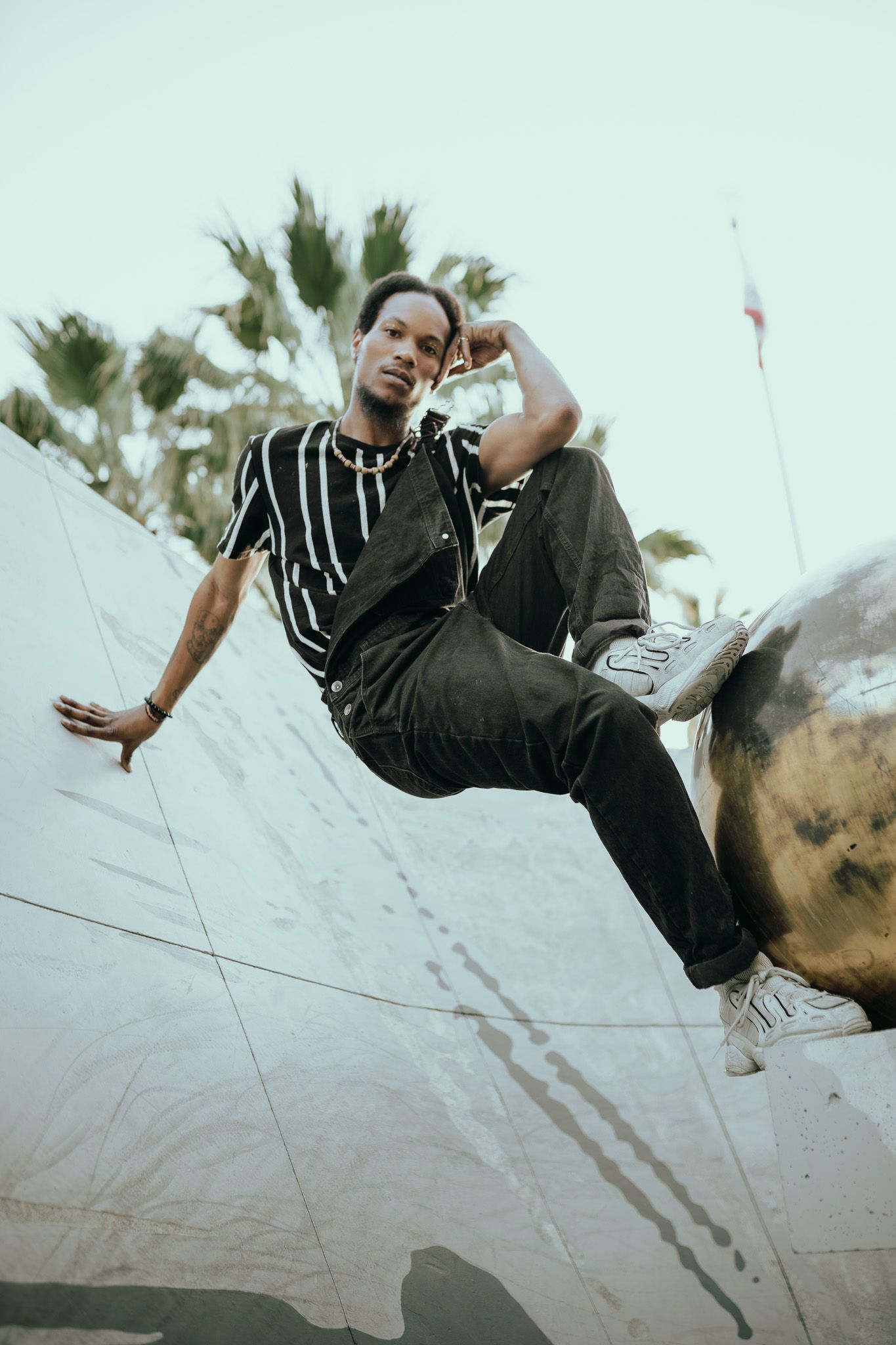 A person in a striped black shirt and pants poses on an angled concrete wall at sunset with palm trees behind them.