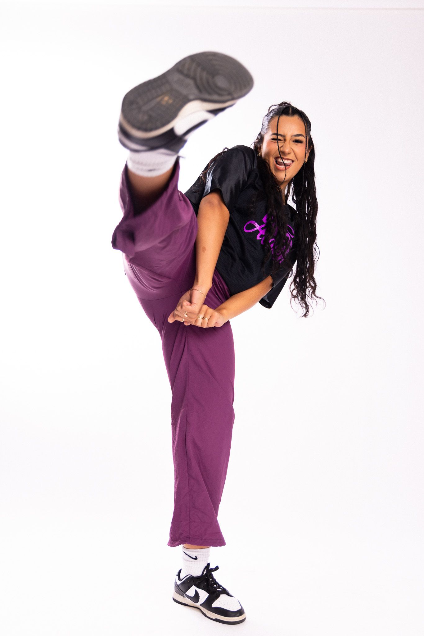 A dancer in purple pants and black top performs an energetic dance move bending forward against a white backdrop.