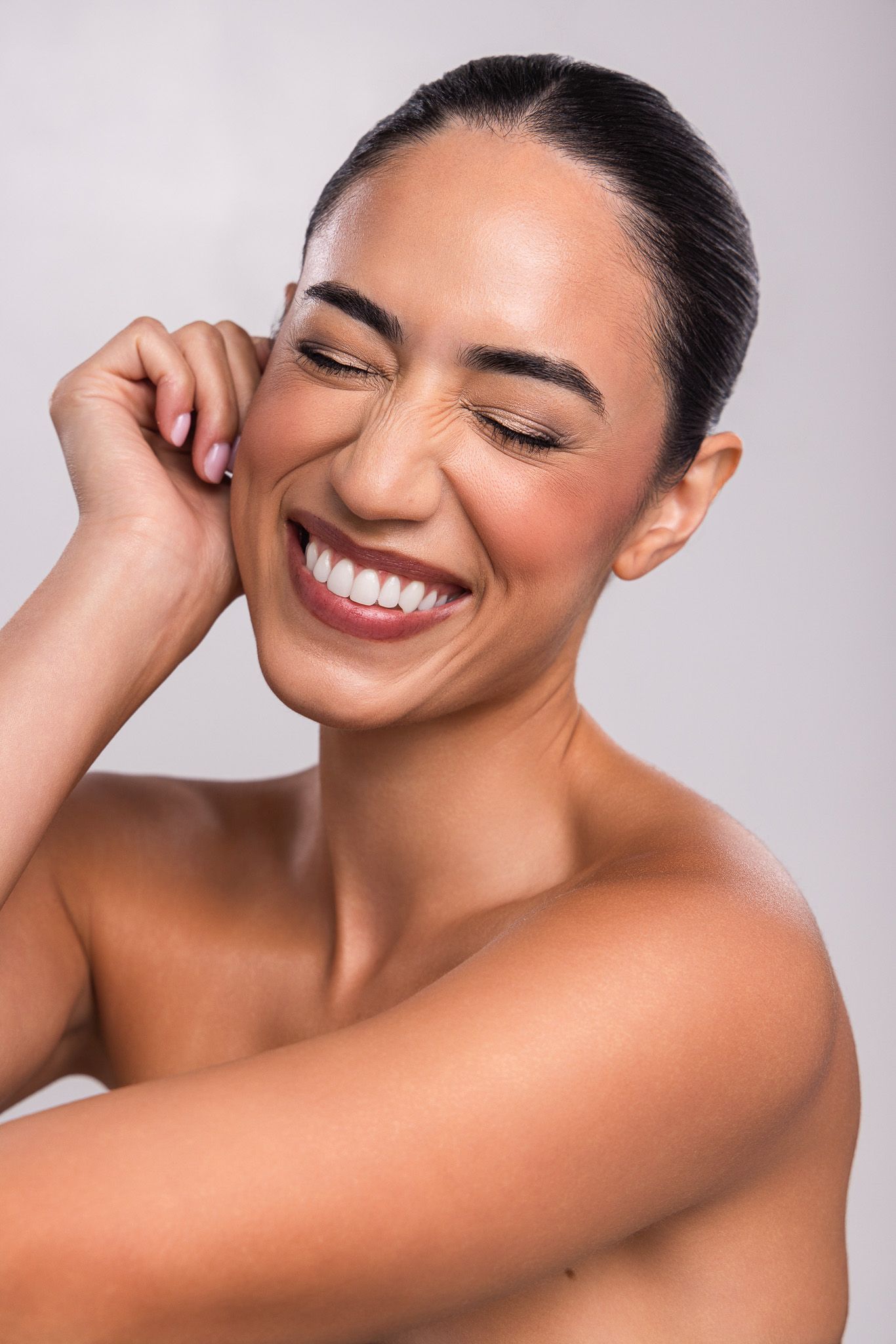 Smiling woman with glowing skin, eyes closed, touching her face against a light gray background.