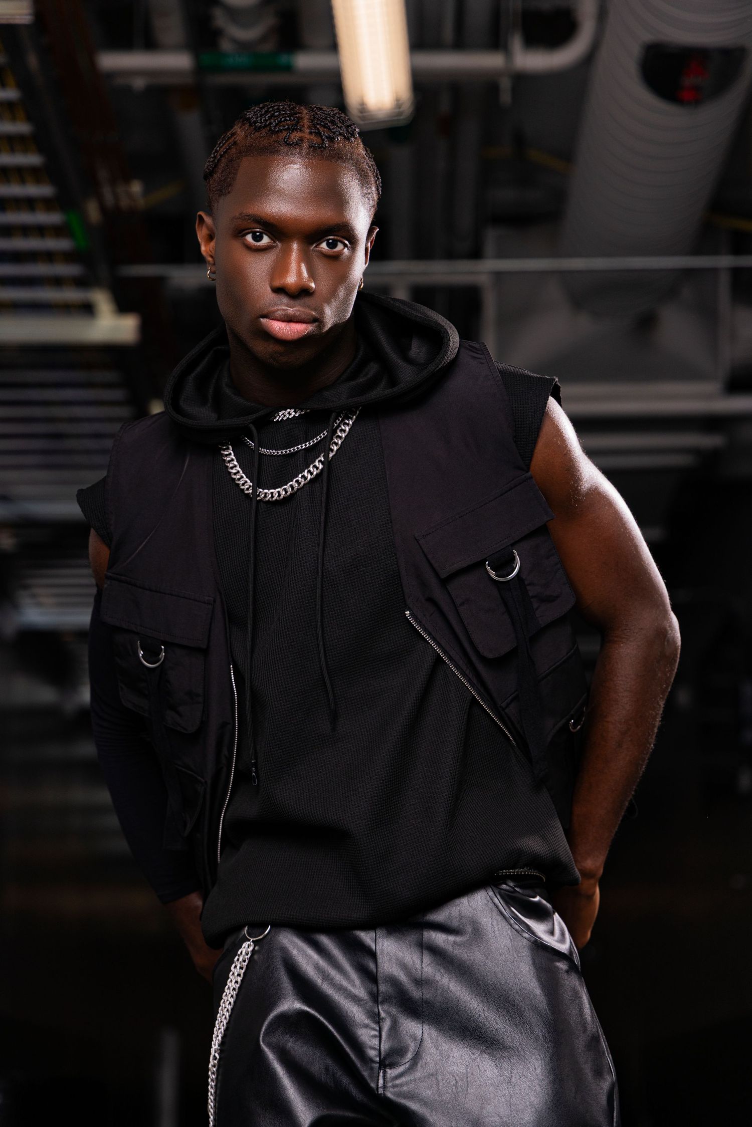 Young Black man in black sleeveless hoodie and chain necklaces poses confidently in dark industrial setting.