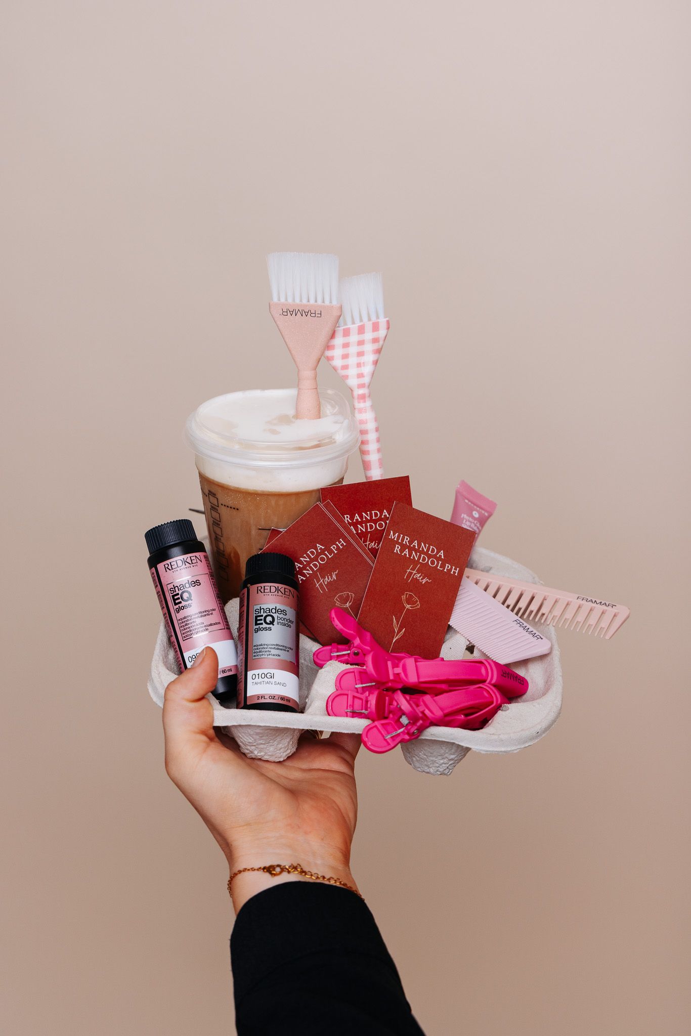 Hand holding a tray with pink hair tools, Redken products, Miranda Kerr booklets, and an iced coffee drink.