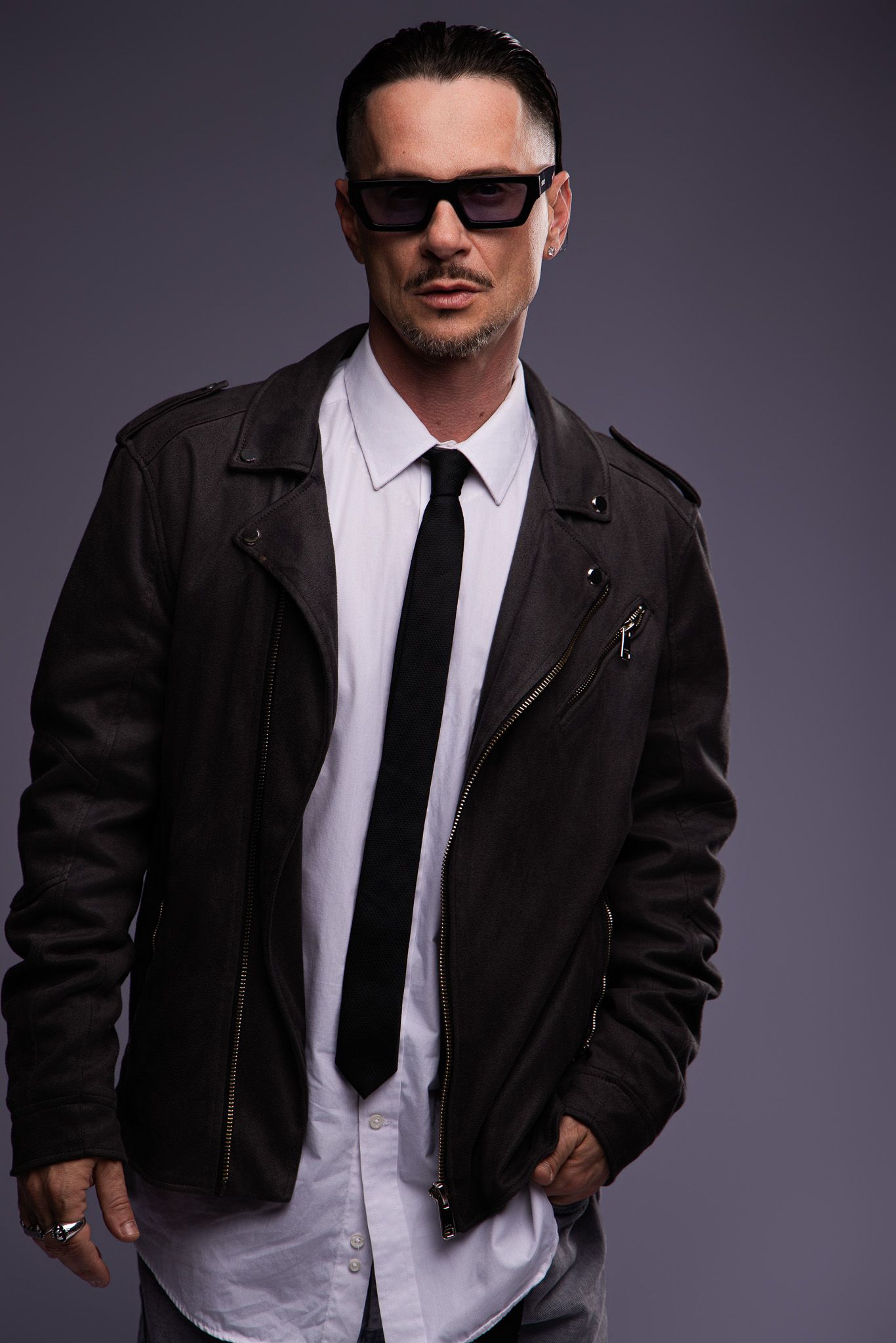 Man in black leather jacket, white shirt, tie, and sunglasses posing confidently against a gray background.