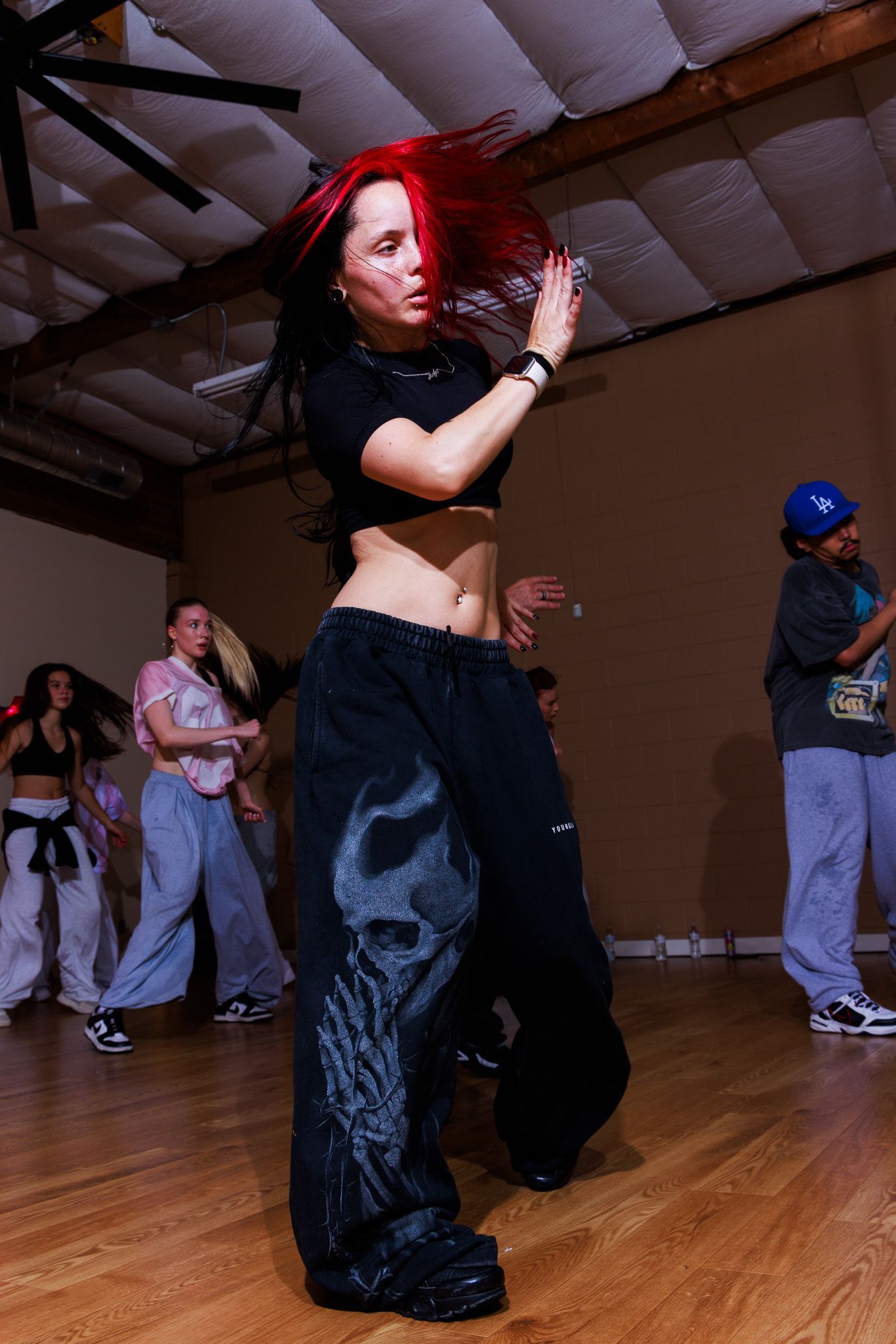 A woman with red hair dances in black baggy pants and crop top in a dance studio with others in the background.