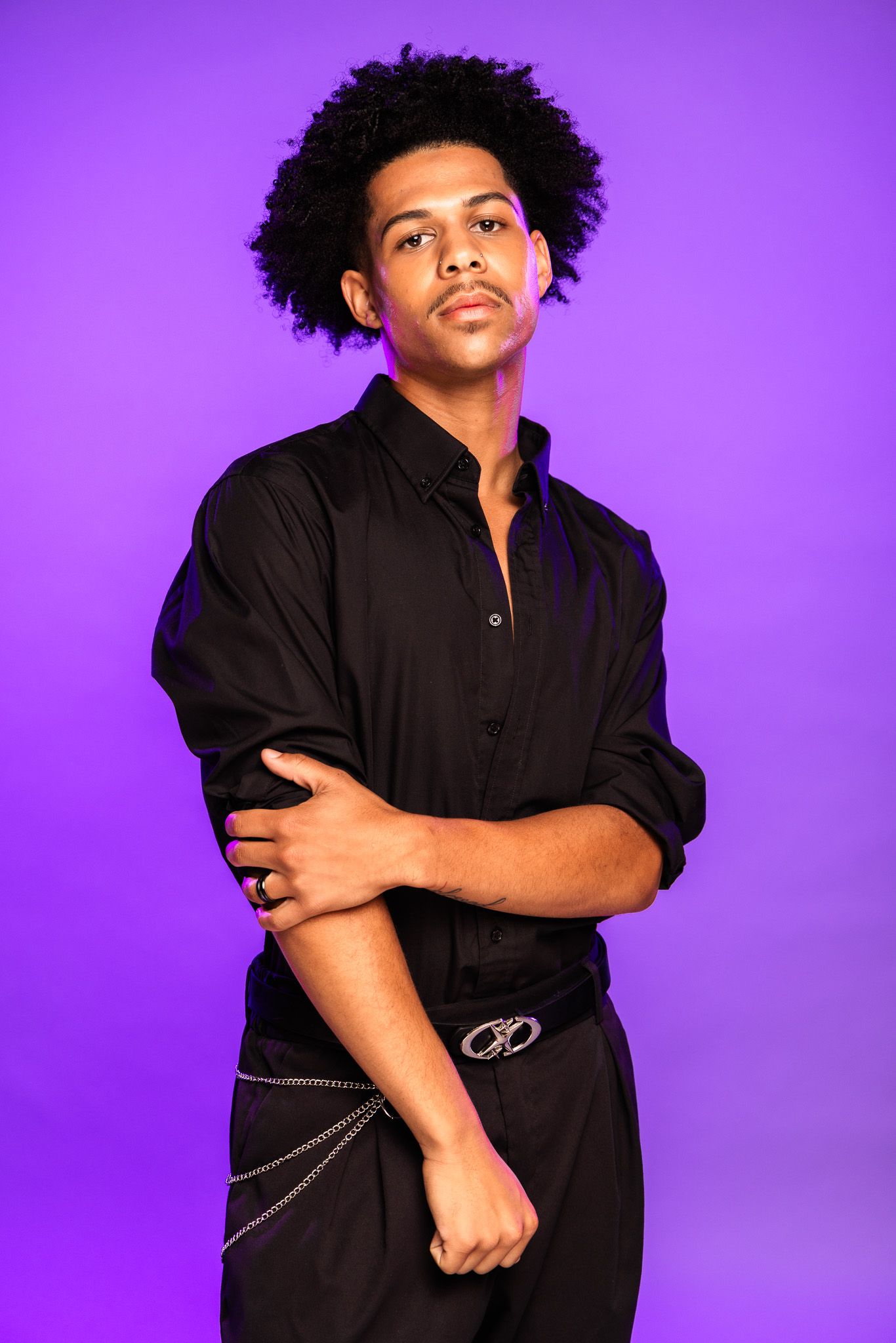 Young Black man with afro in black shirt and chain belt posing confidently against purple background.