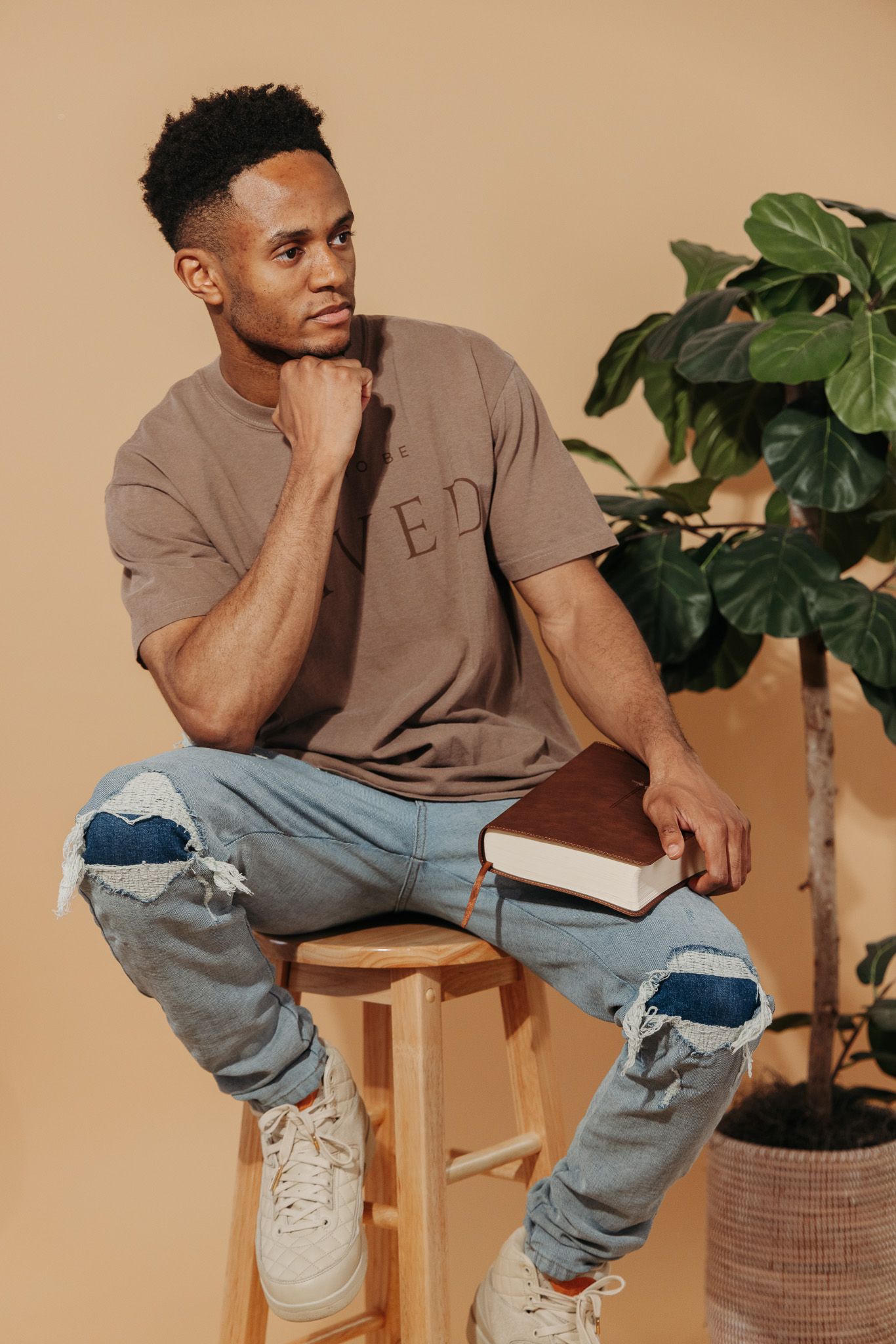 Fashion model wearing distressed jeans and brown t-shirt poses on wooden stool against beige backdrop with potted plant.