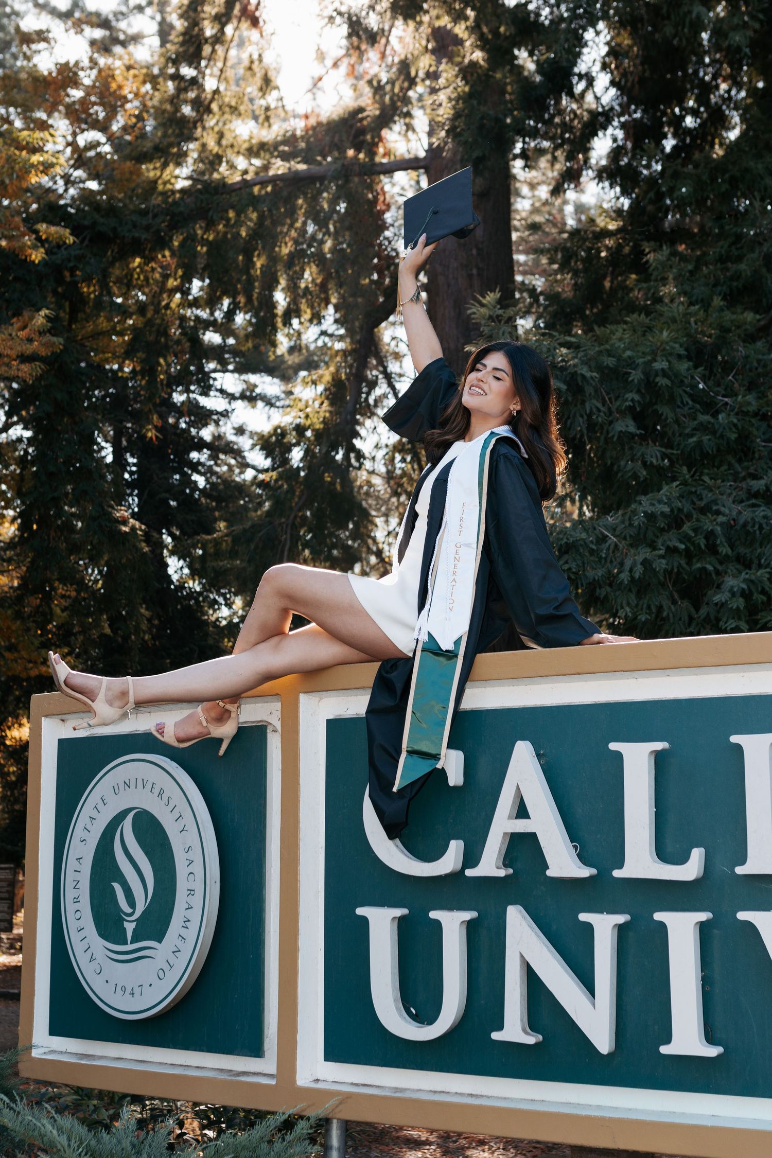 A graduation photoshoot features a creative pose on top of a university sign with letters framed against trees.