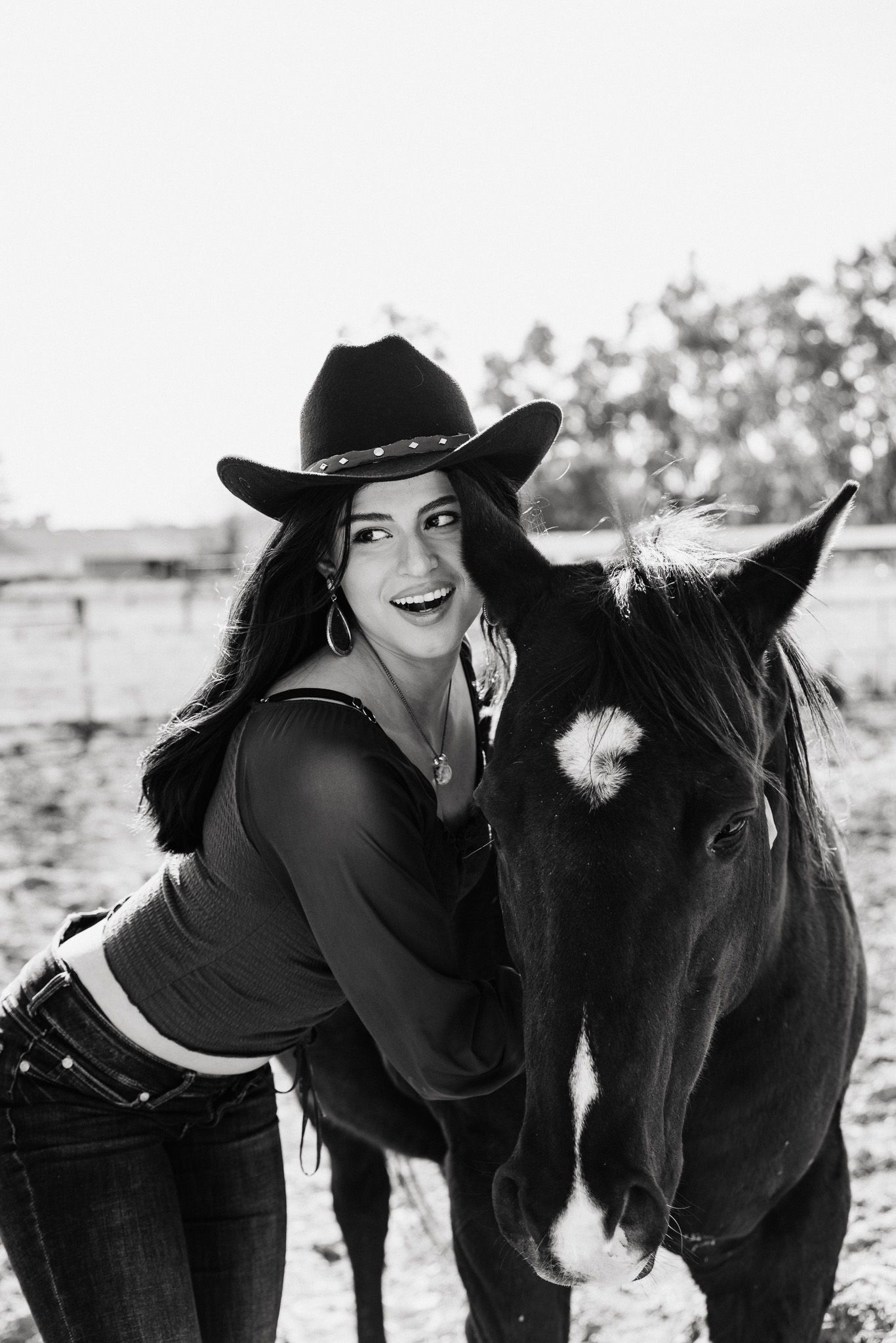 Black and white portrait of equestrian with cowboy hat embracing horse outdoors in countryside setting.