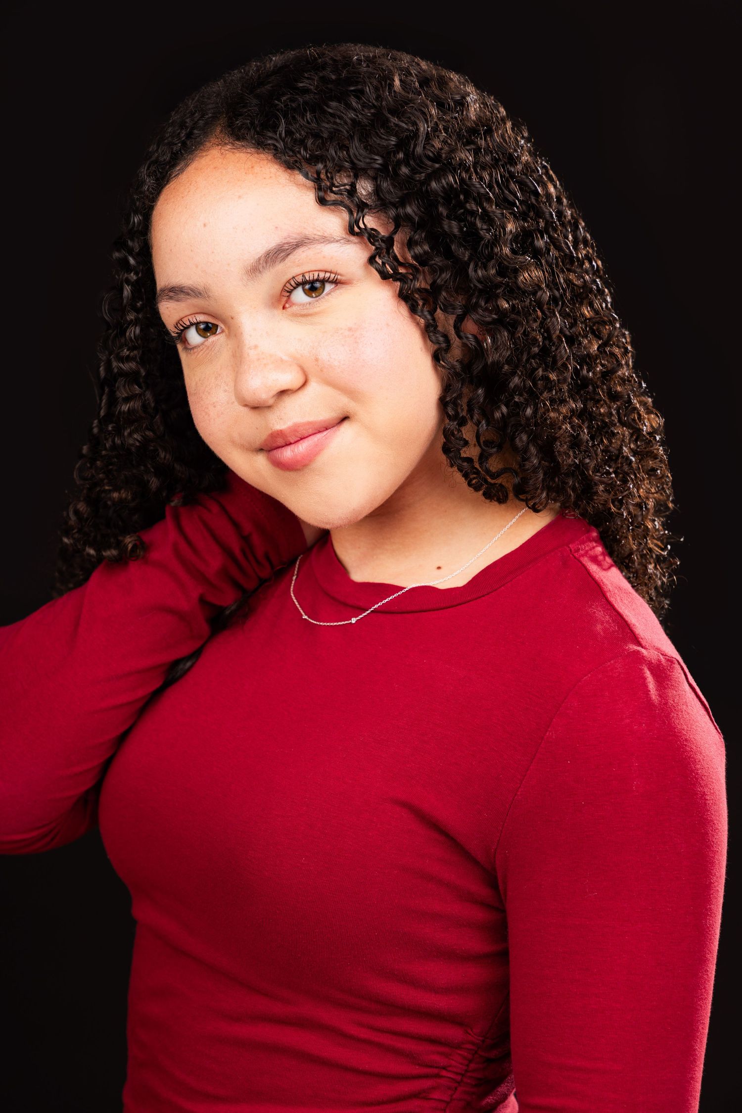 Studio portrait with natural curly hair and red long-sleeve top against dark background.