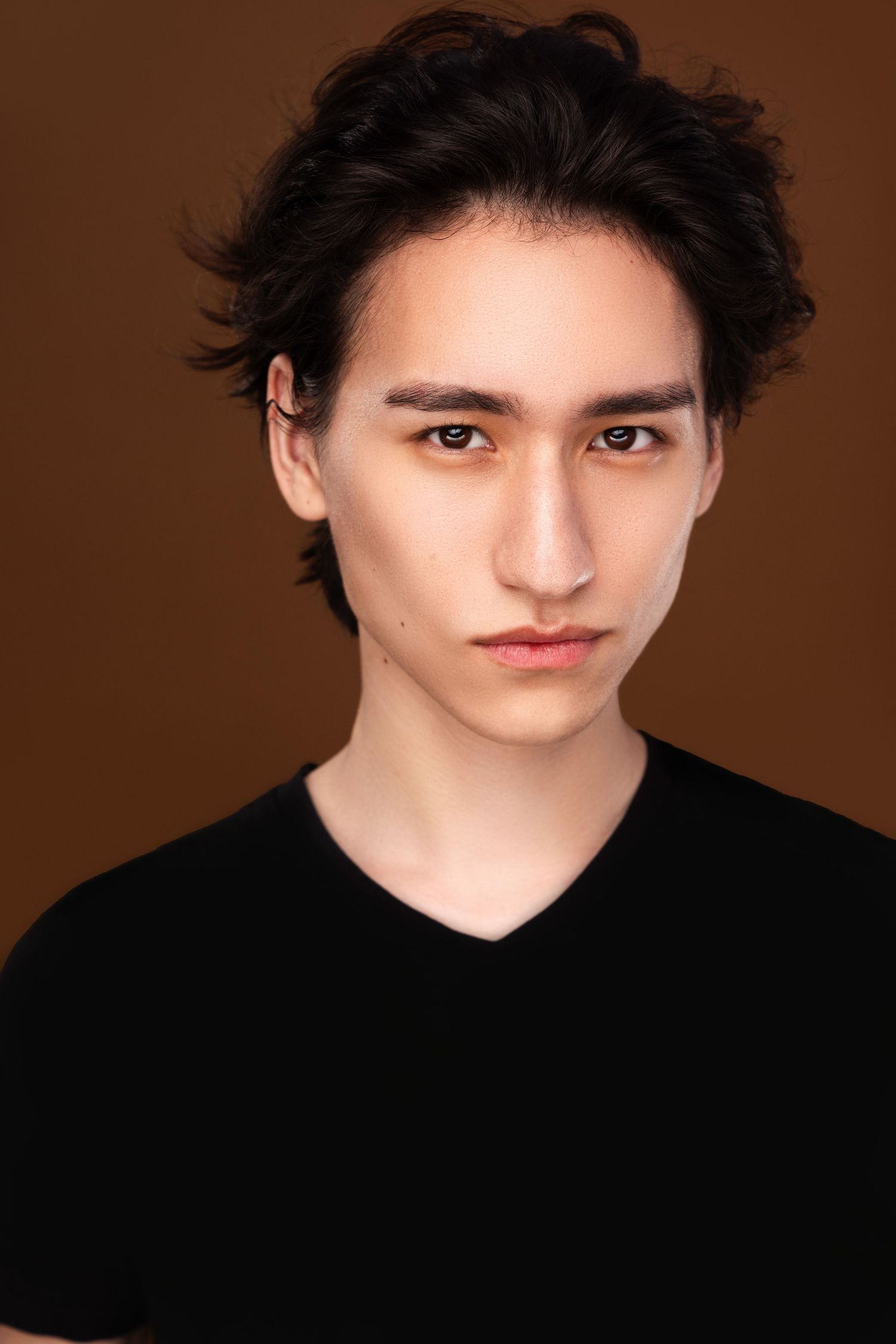 Dramatic close-up portraits against brown background wearing black top with tousled dark hair.