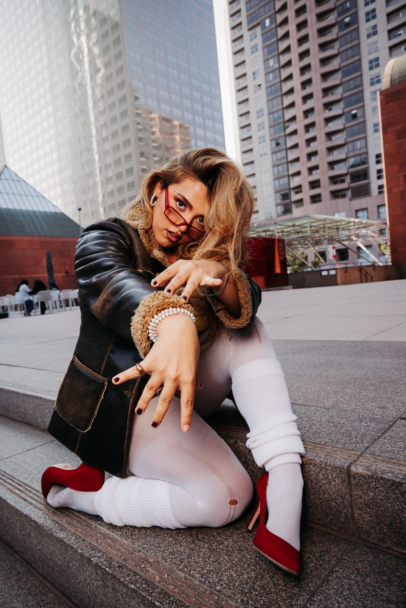 Urban fashion portrait with leather jacket, white pants and red heels against city skyscraper backdrop.