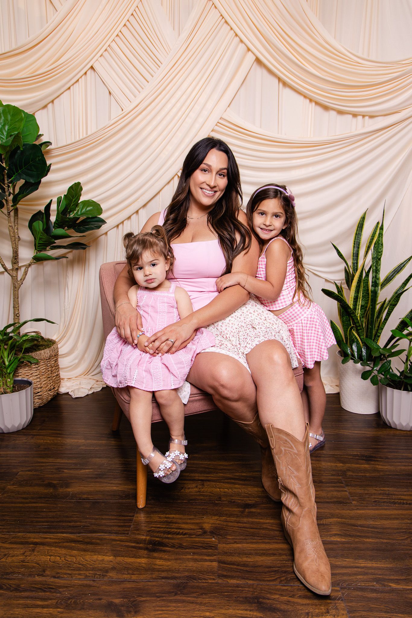 A woman in a pink dress sits with two young girls in matching pink outfits, surrounded by plants and draped fabric backdrop.