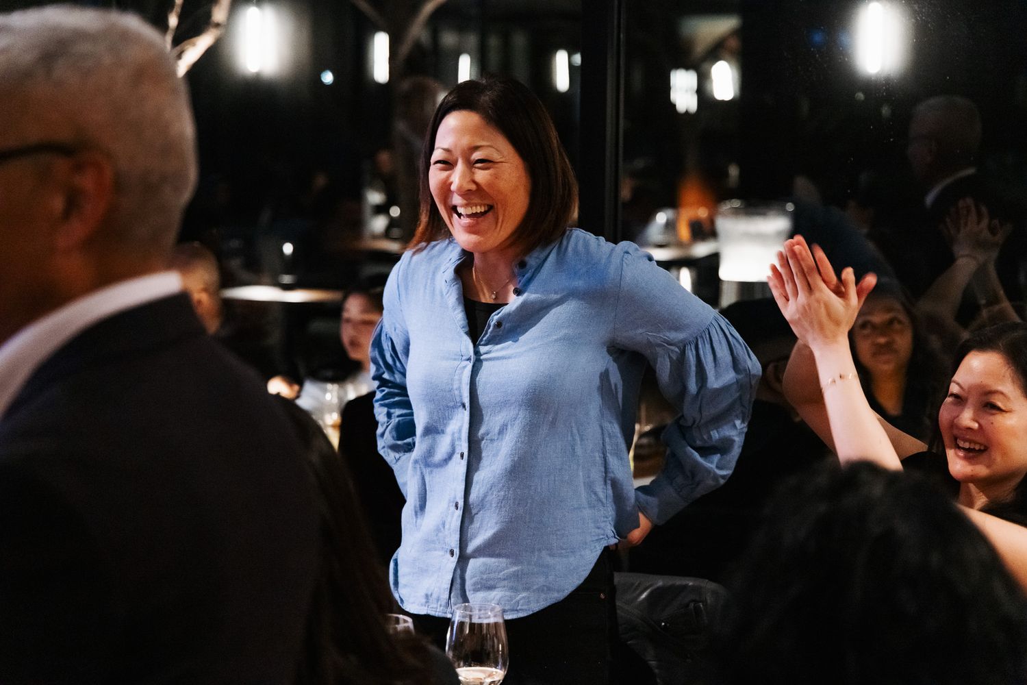 A person in a light blue button-down shirt laughs joyfully during a nighttime gathering with others.
