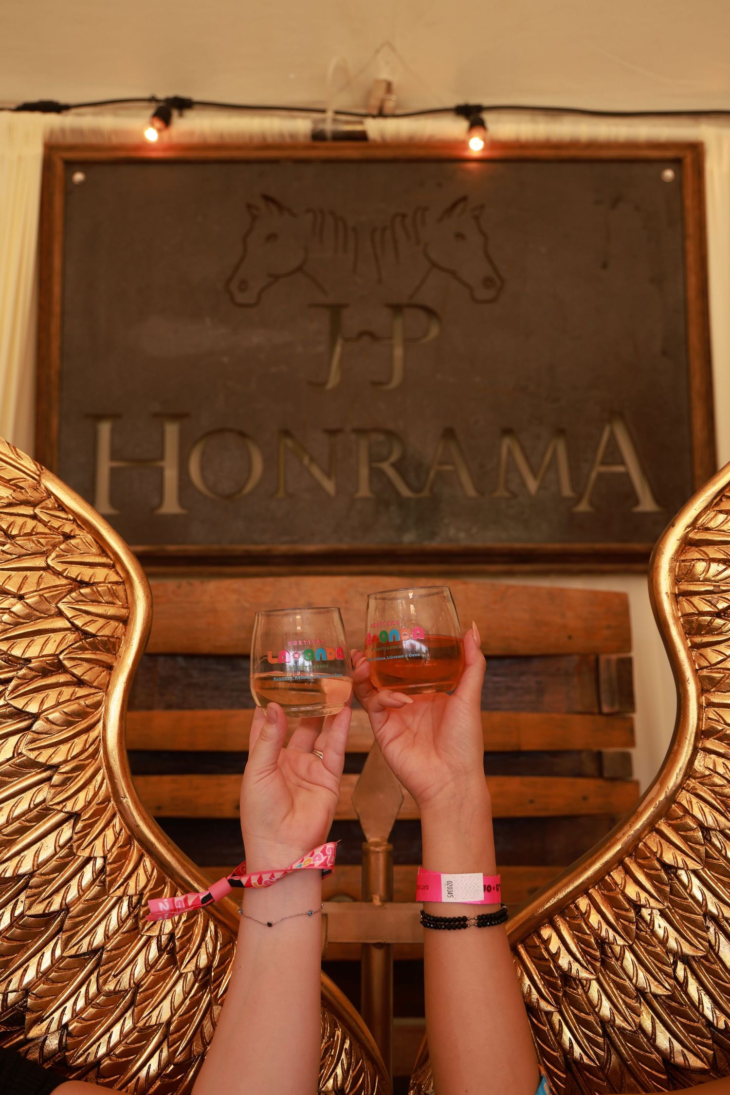 Two hands holding wine glasses toast beneath a rustic Honrama sign with decorative golden wings in the background.