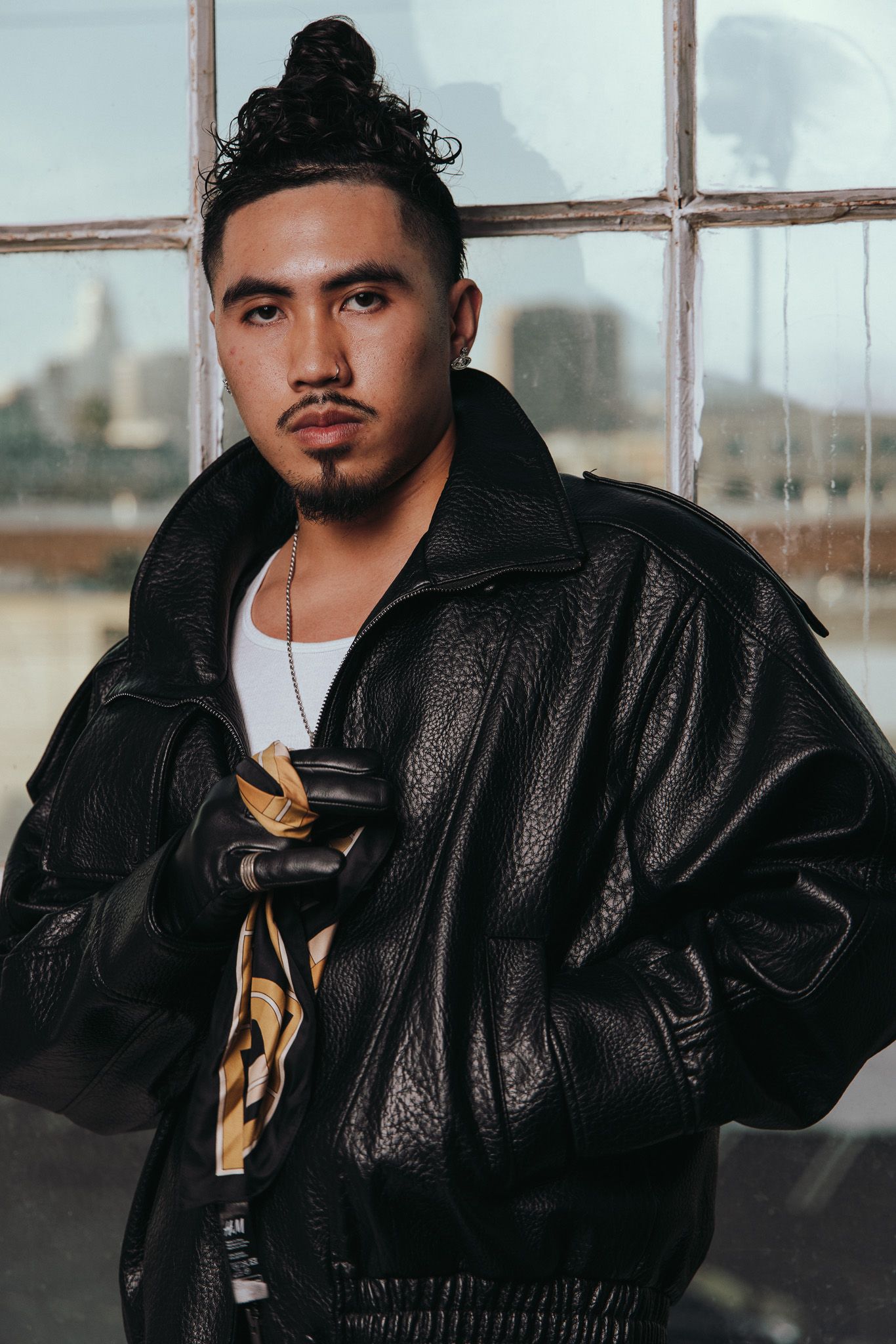 Urban fashion portrait in a black leather jacket and white tank top near a window with natural lighting.