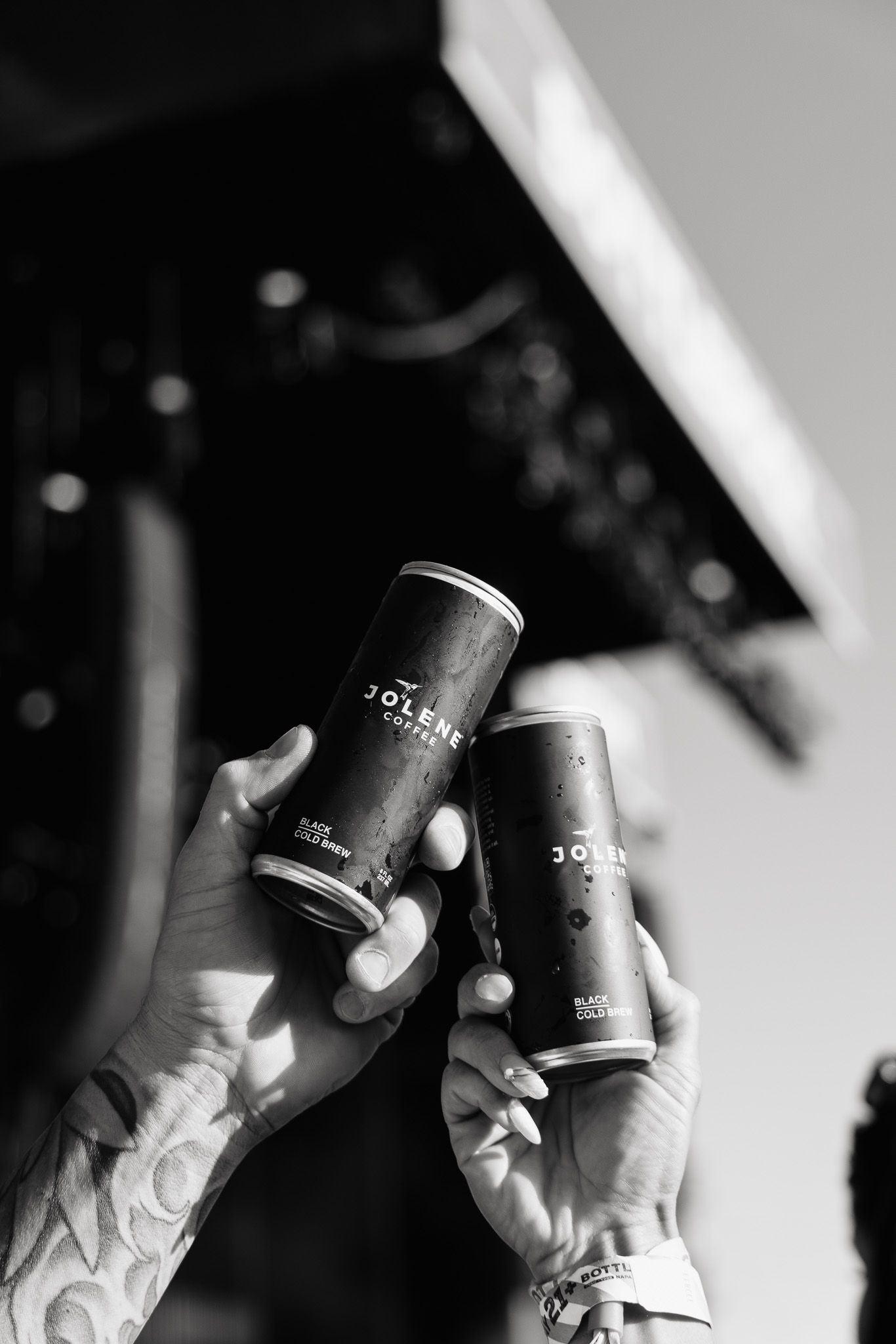 Two tattooed hands hold up dark beverage cans in a celebratory cheers gesture against a black and white background.