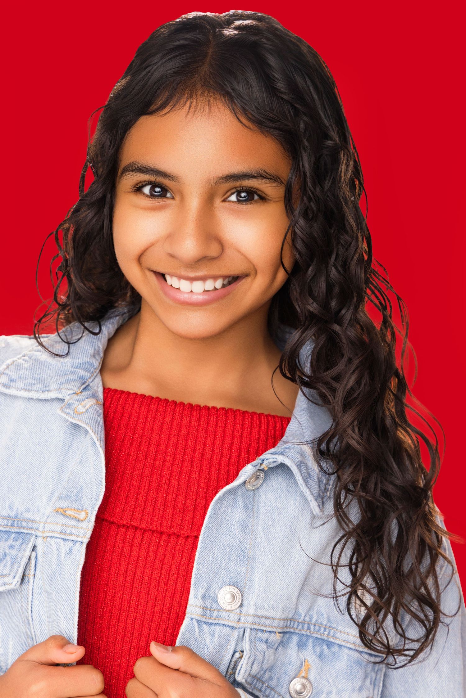 Woman with curly dark hair wearing a red sweater and denim jacket poses against vibrant red background.