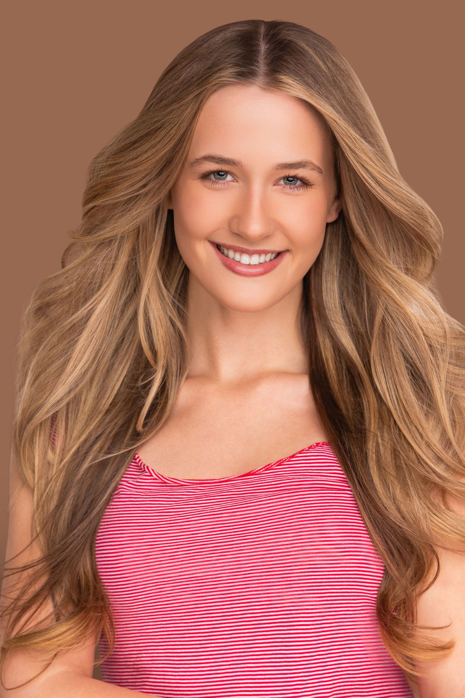 Blonde woman with wavy hair wearing a pink and white striped sleeveless top smiles at camera.