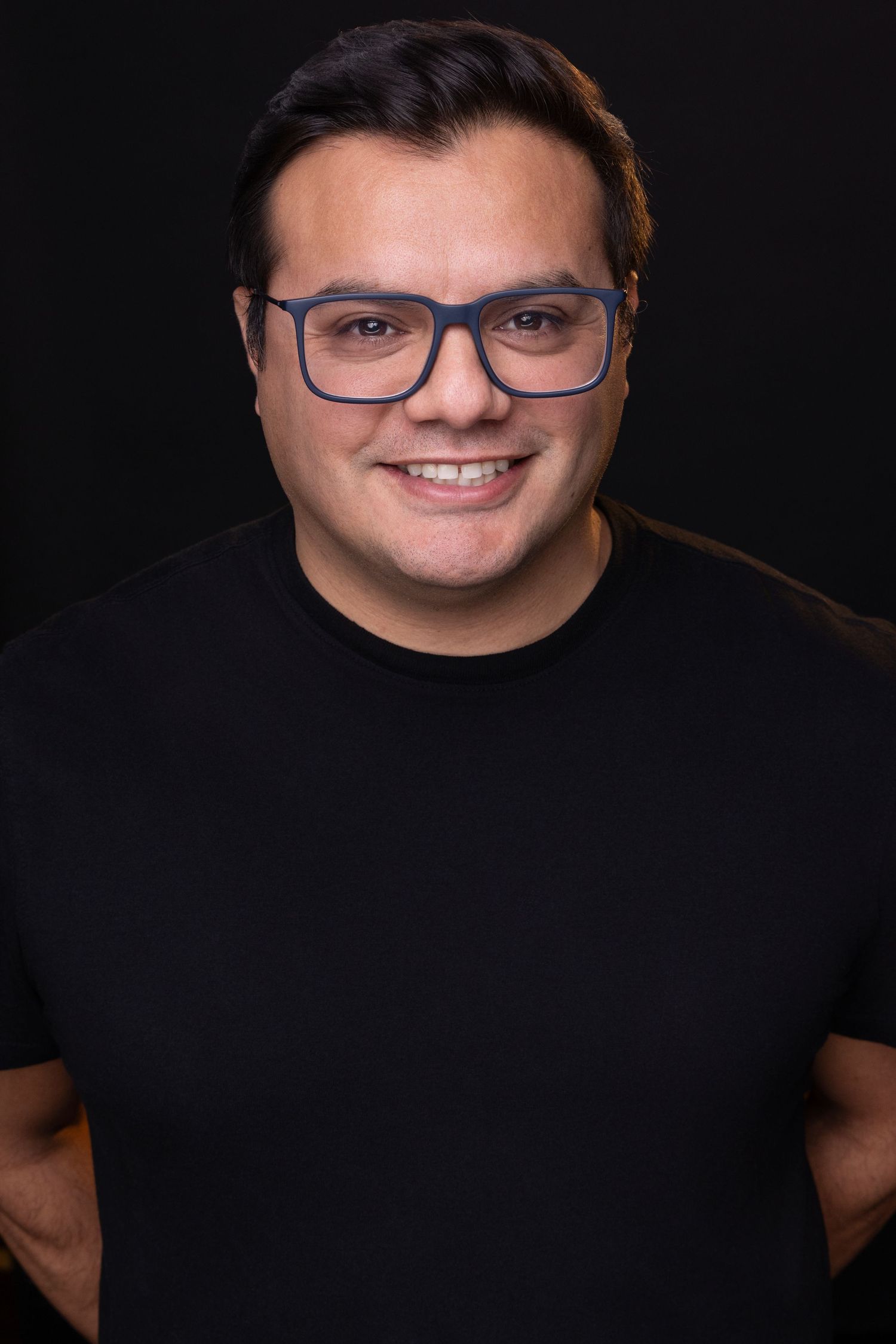 Professional headshot of someone wearing glasses and a black t-shirt against a dark background with a warm smile.