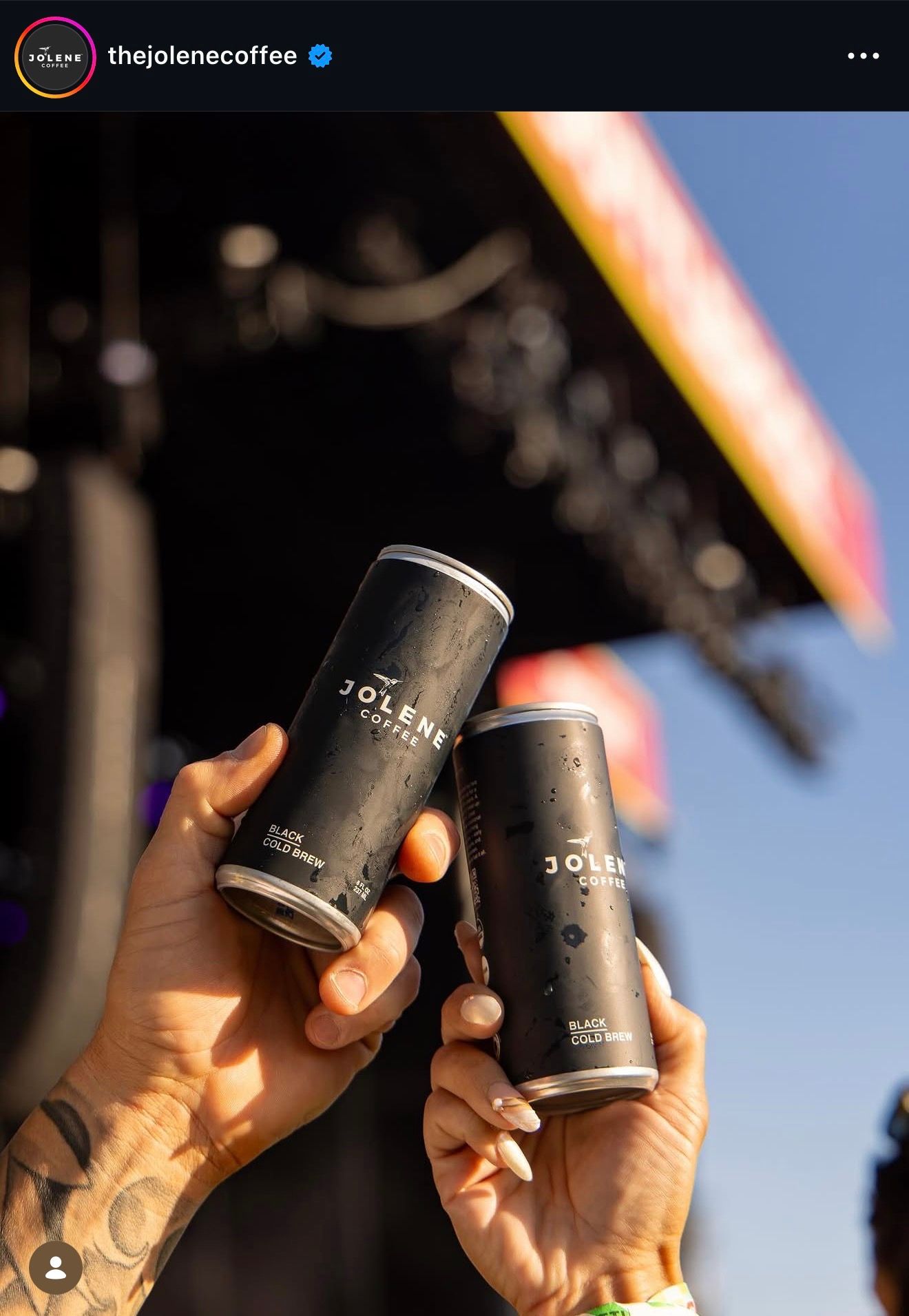 Two hands holding Jolene black cold brew coffee cans at an outdoor festival with a stage in the background.