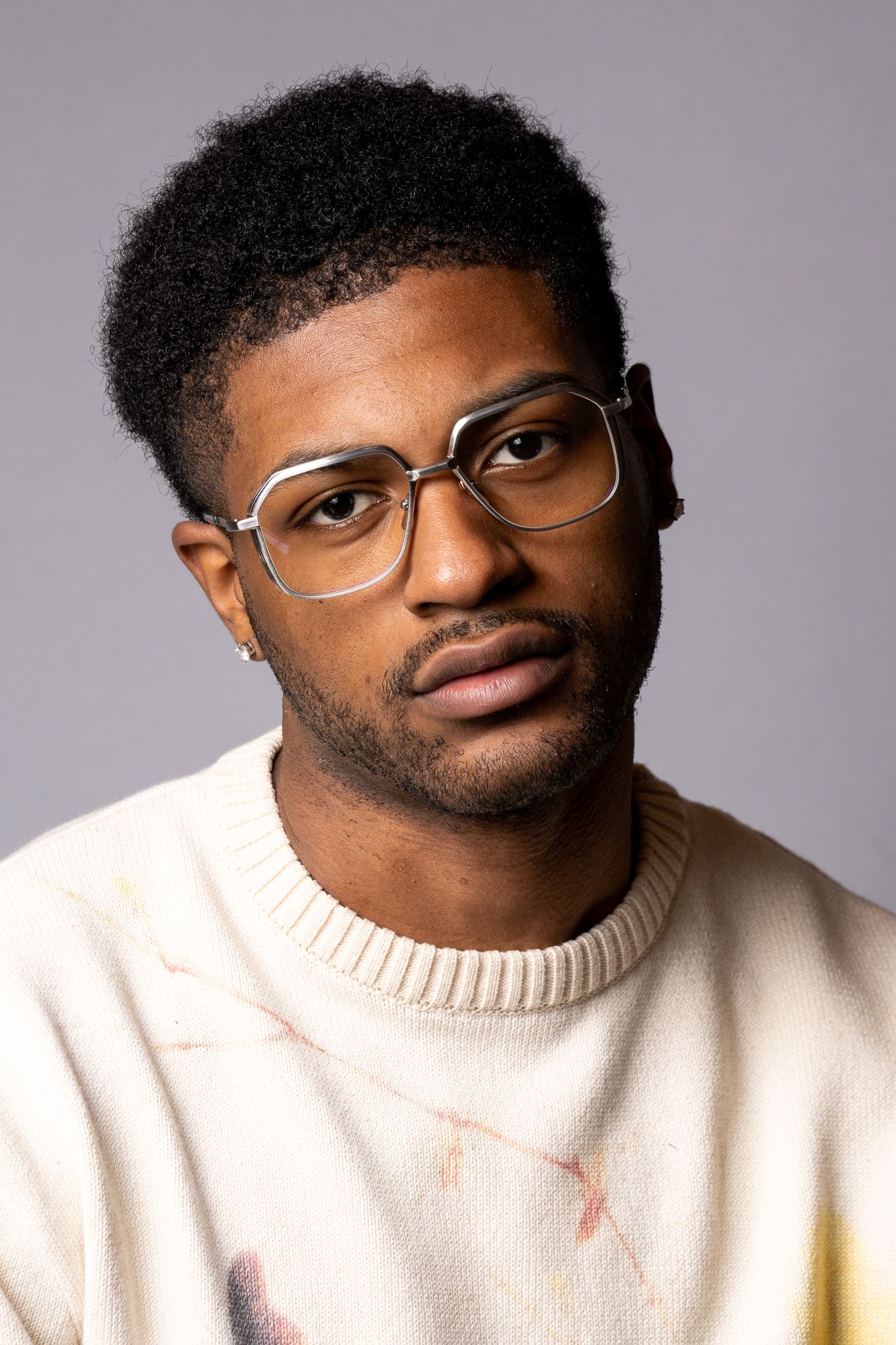 Young Black man with curly hair wearing large square glasses and cream sweater poses against gray background.