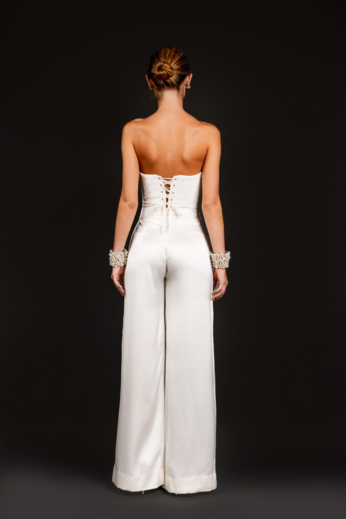 Back view of a white strapless corset lace-up satin wide-leg jumpsuit with crystal cuff bracelets.