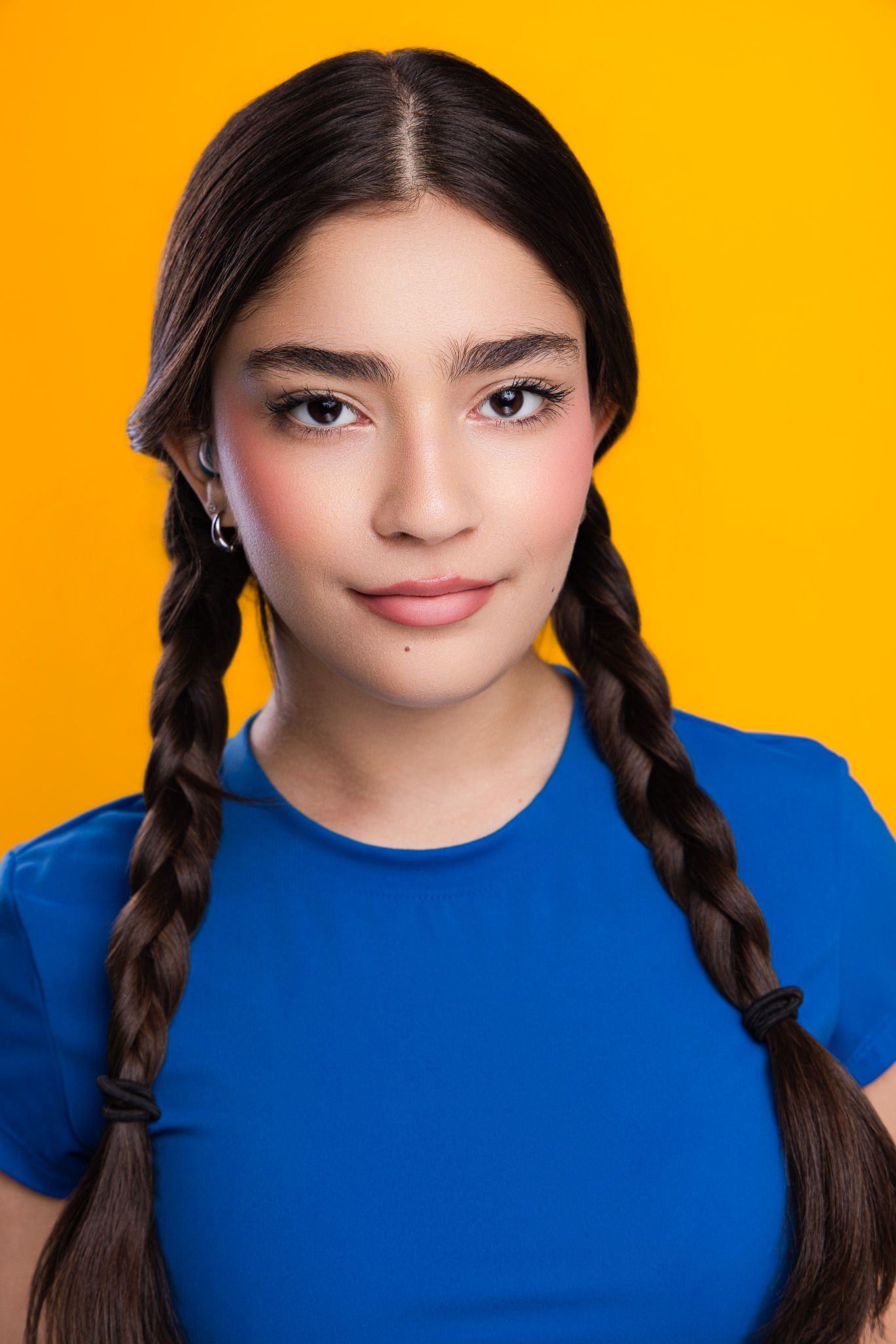 Young woman with dark braids wearing a blue t-shirt poses against a bright yellow background.