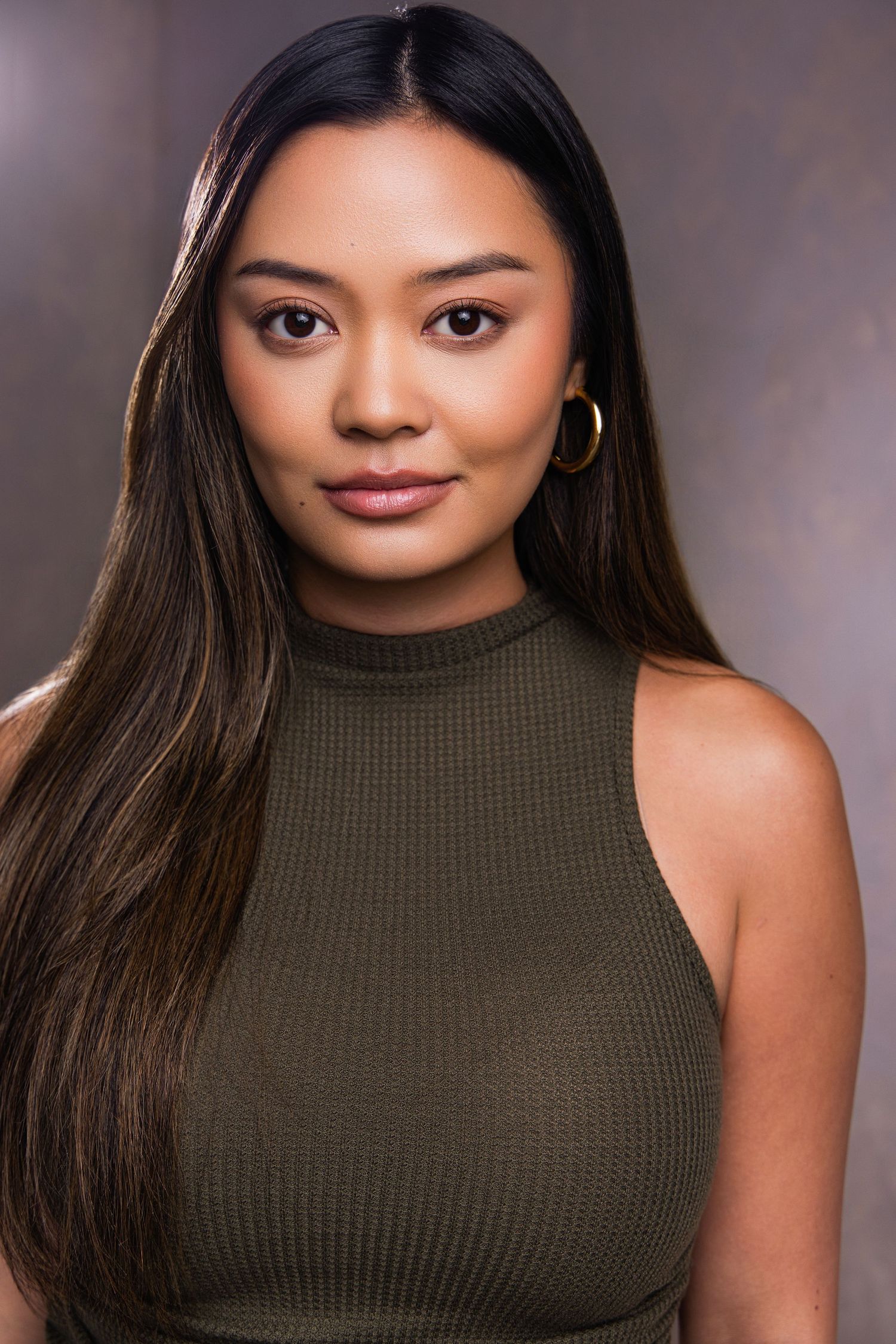 Young woman with long dark hair wearing olive green sleeveless top and gold hoop earrings poses for professional headshot.