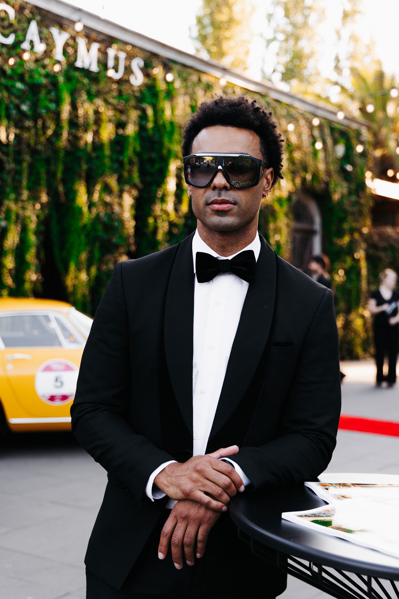 A person in a black tuxedo and sunglasses poses confidently against a natural outdoor background.
