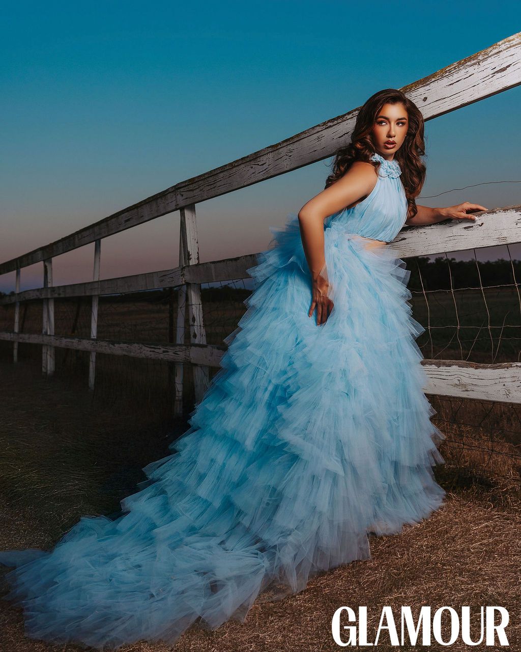 A flowing light blue tulle gown creates an ethereal silhouette against a rustic wooden fence at sunset.
