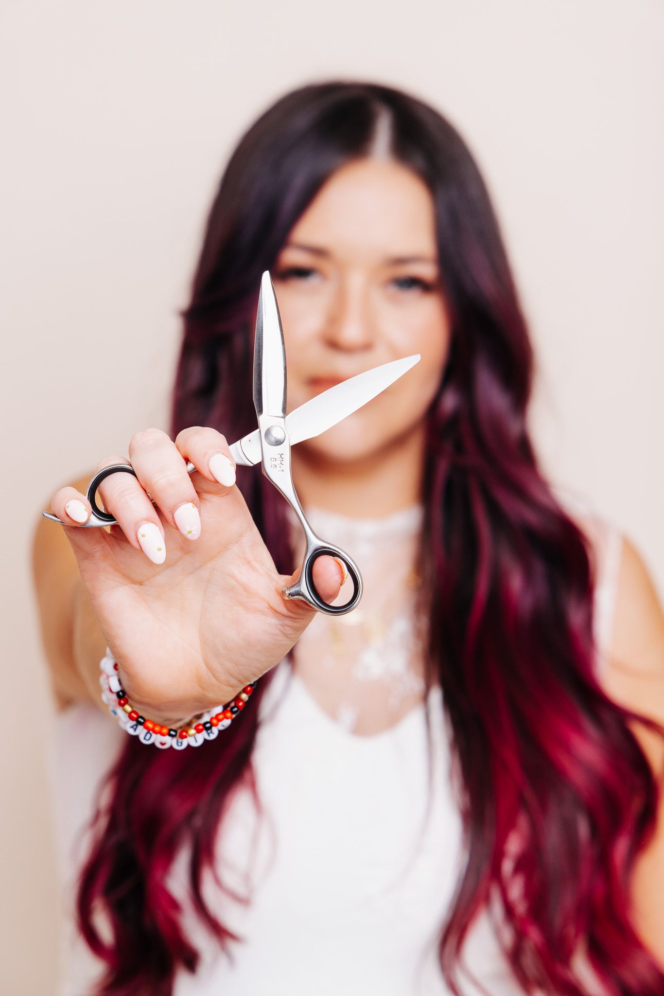 Woman with long burgundy hair holding professional hair scissors toward camera in a salon setting.