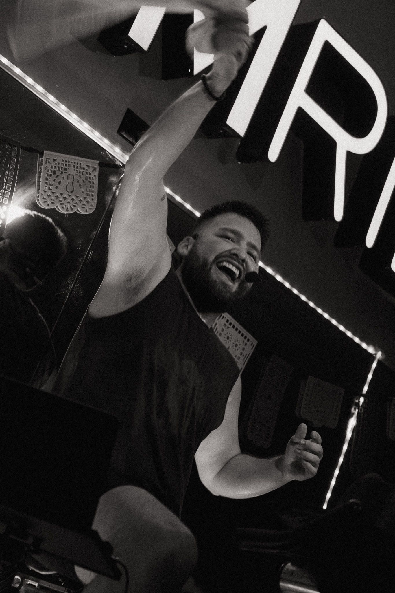 Black and white photo of a bearded man laughing and raising his arm at a lively music venue.