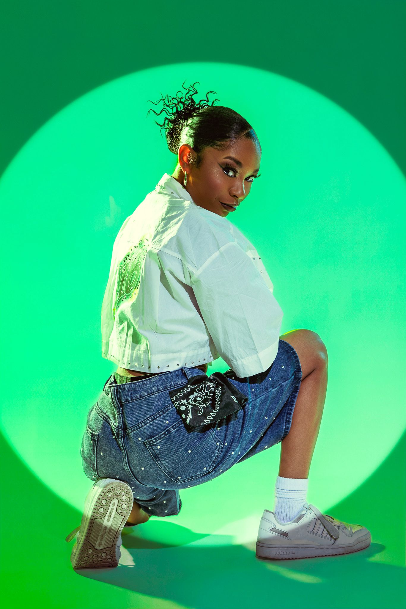 Young woman in white crop jacket and denim skirt poses confidently against vibrant green circular backdrop.