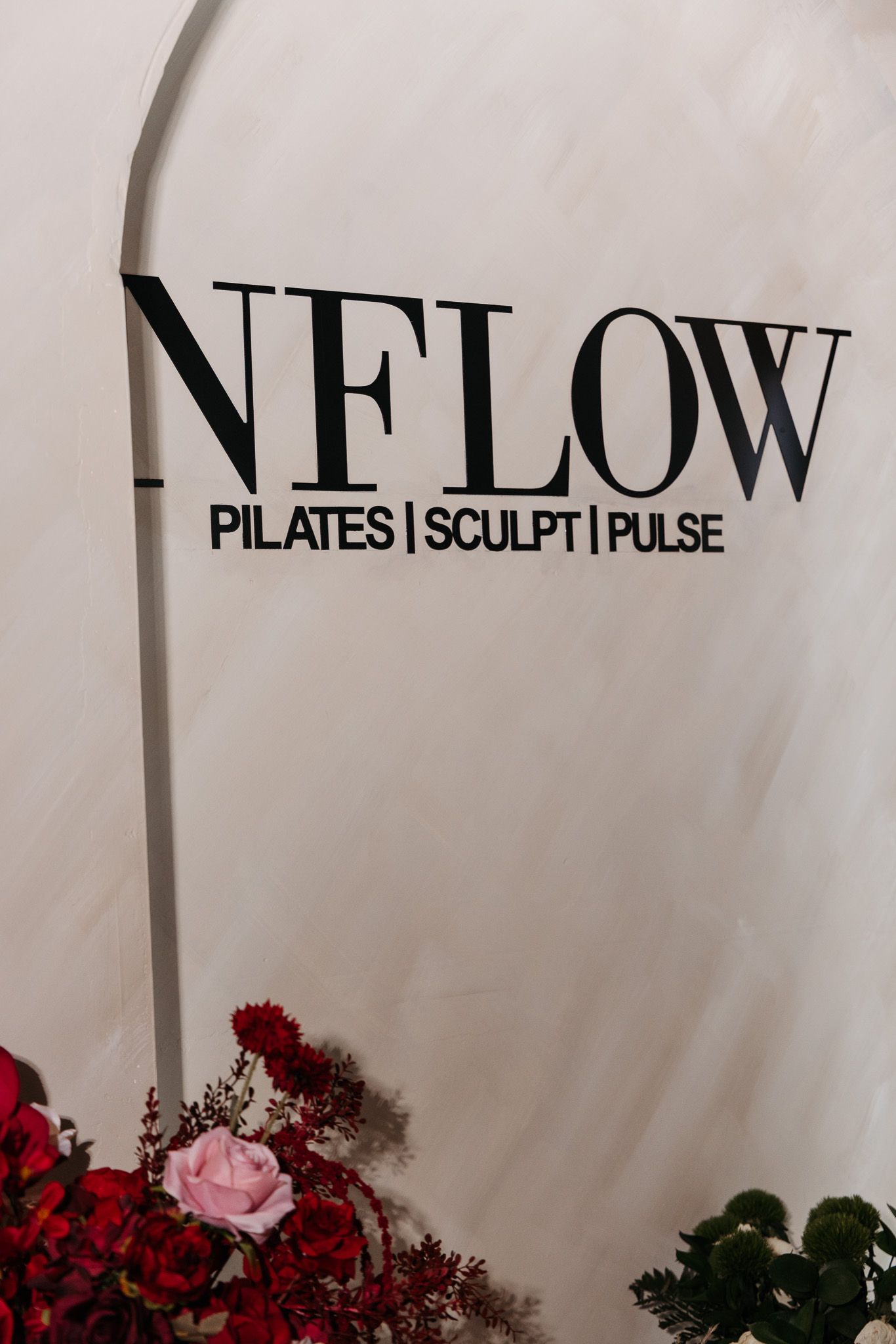 Black NFLOW Pilates studio logo on white wall with red and pink floral decorations.