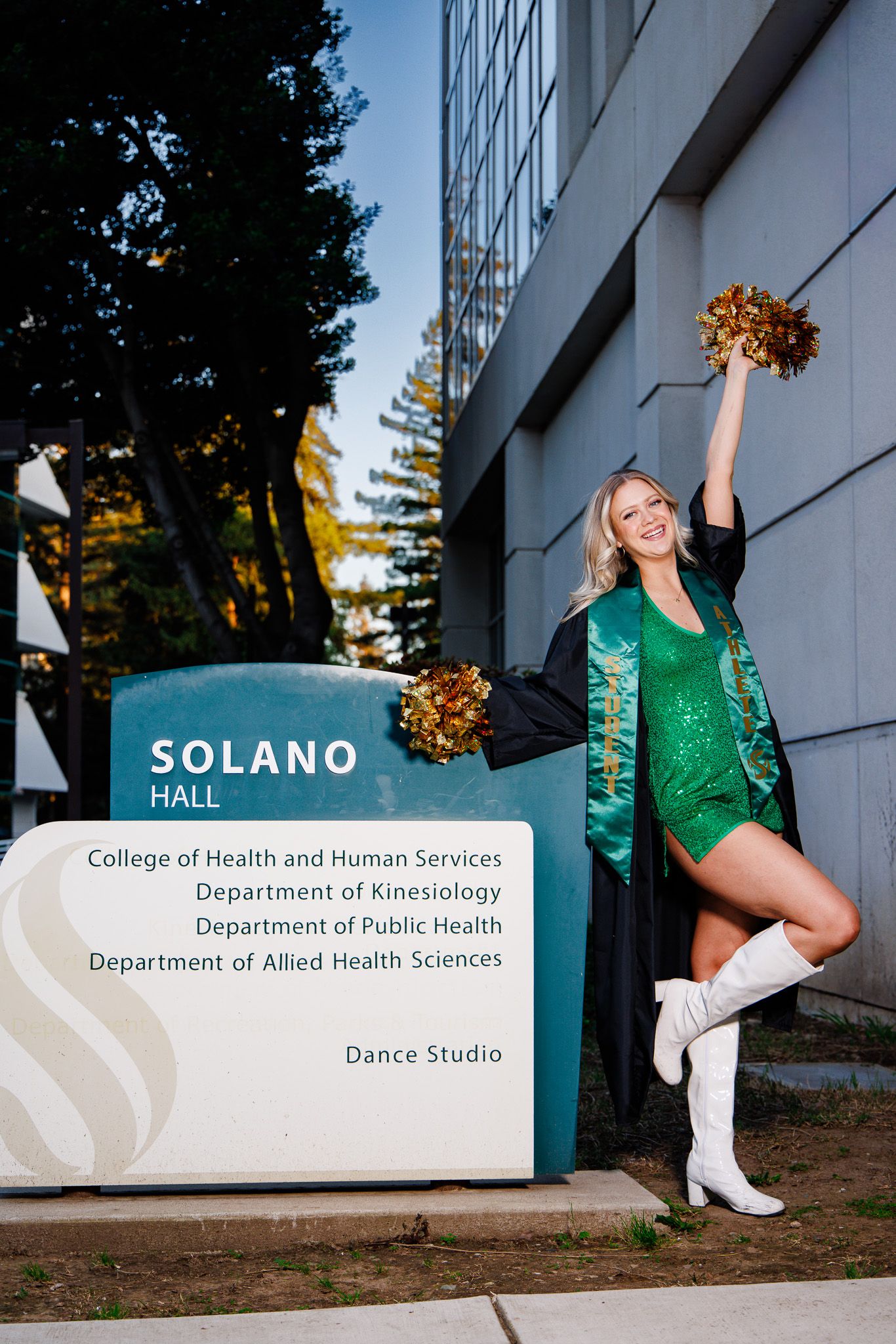 Cheerleader in green sequin outfit and graduation stole poses with pom-poms by Solano Hall sign at college campus.