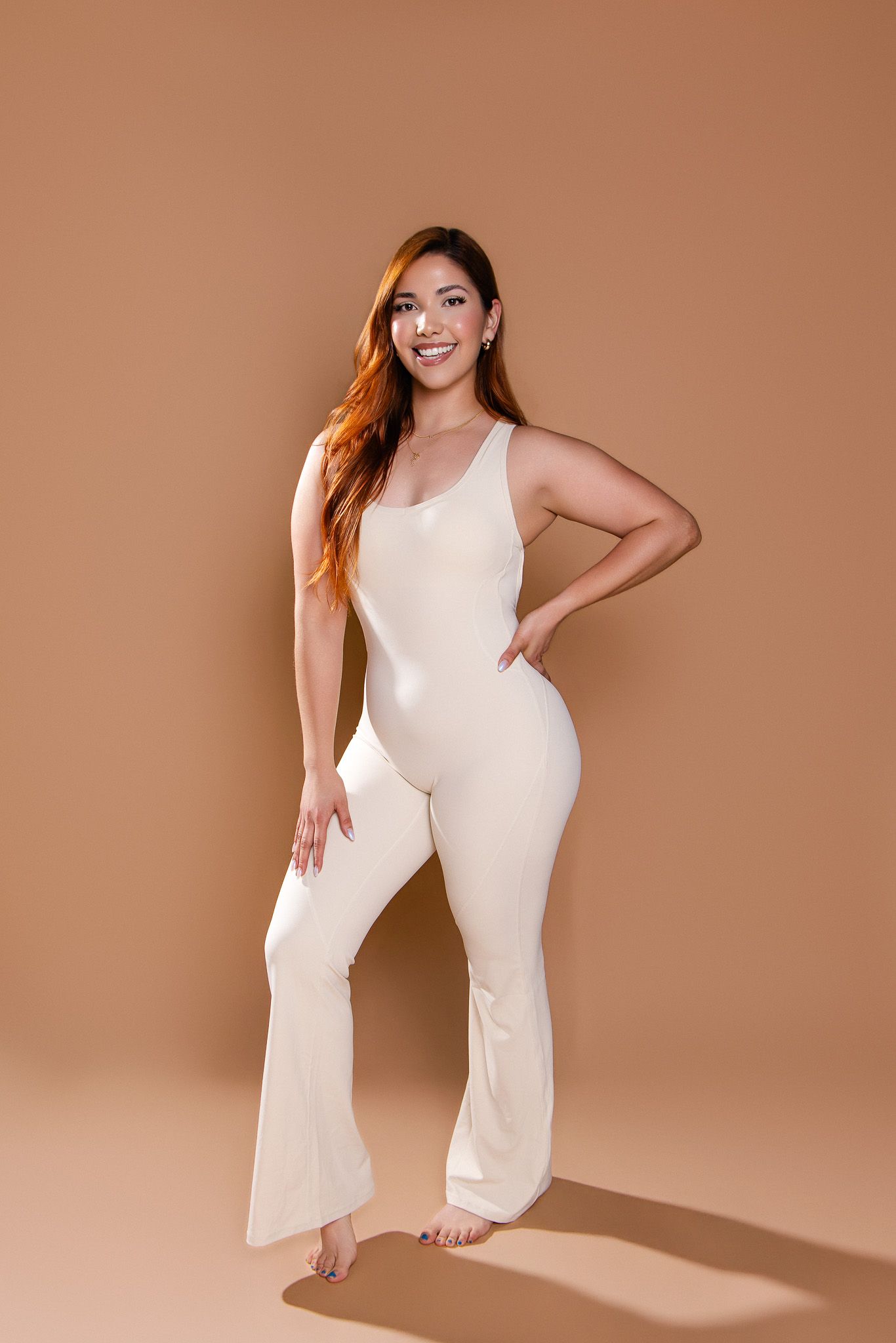Smiling woman in cream flare jumpsuit posing confidently on beige background.