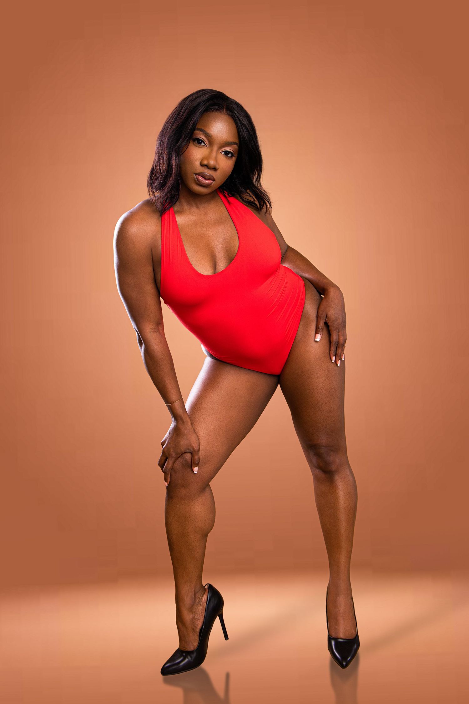 Woman in red swimsuit and black heels posing confidently against a warm brown studio background.