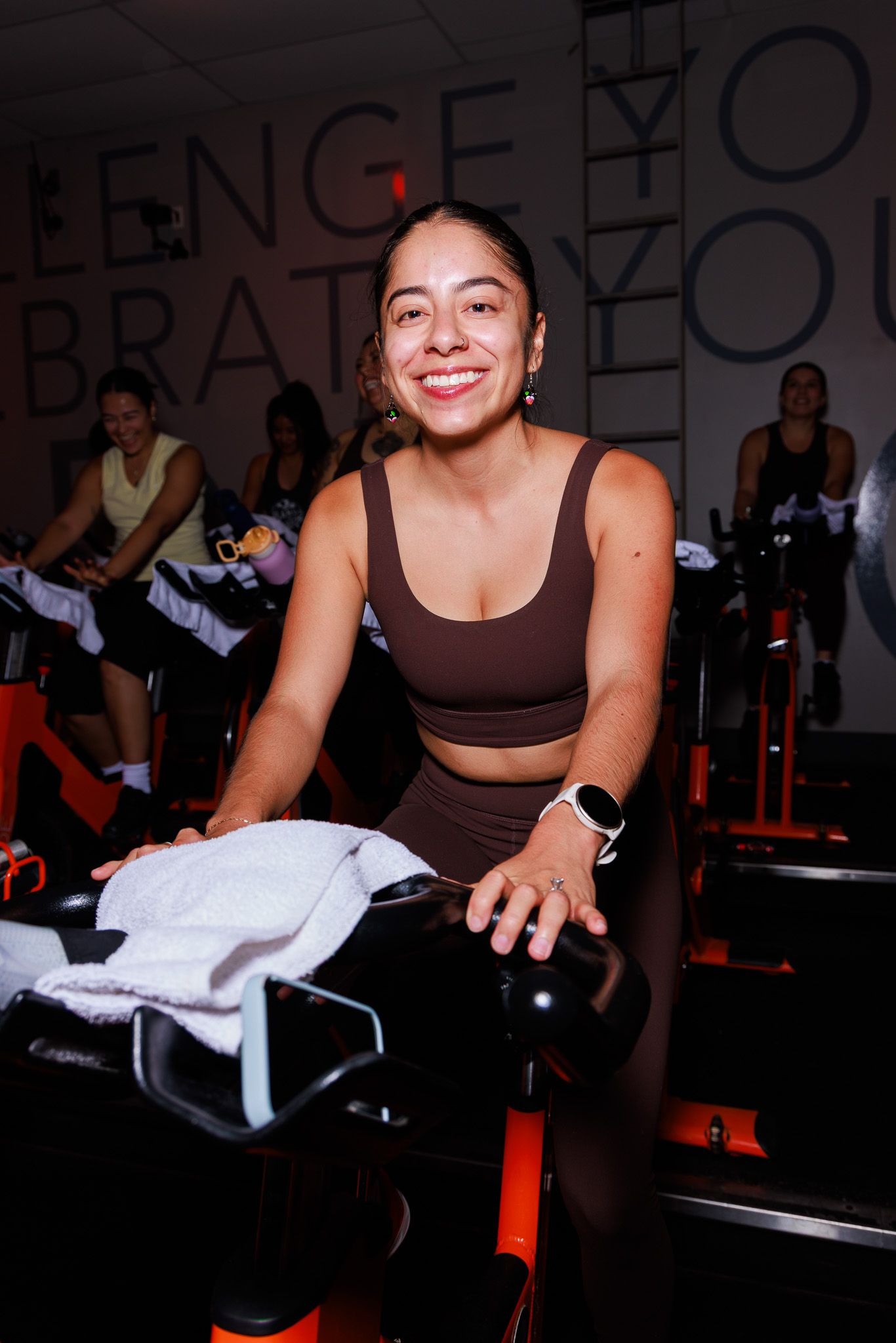Smiling woman in brown athletic wear on a stationary bike at an indoor cycling fitness class.