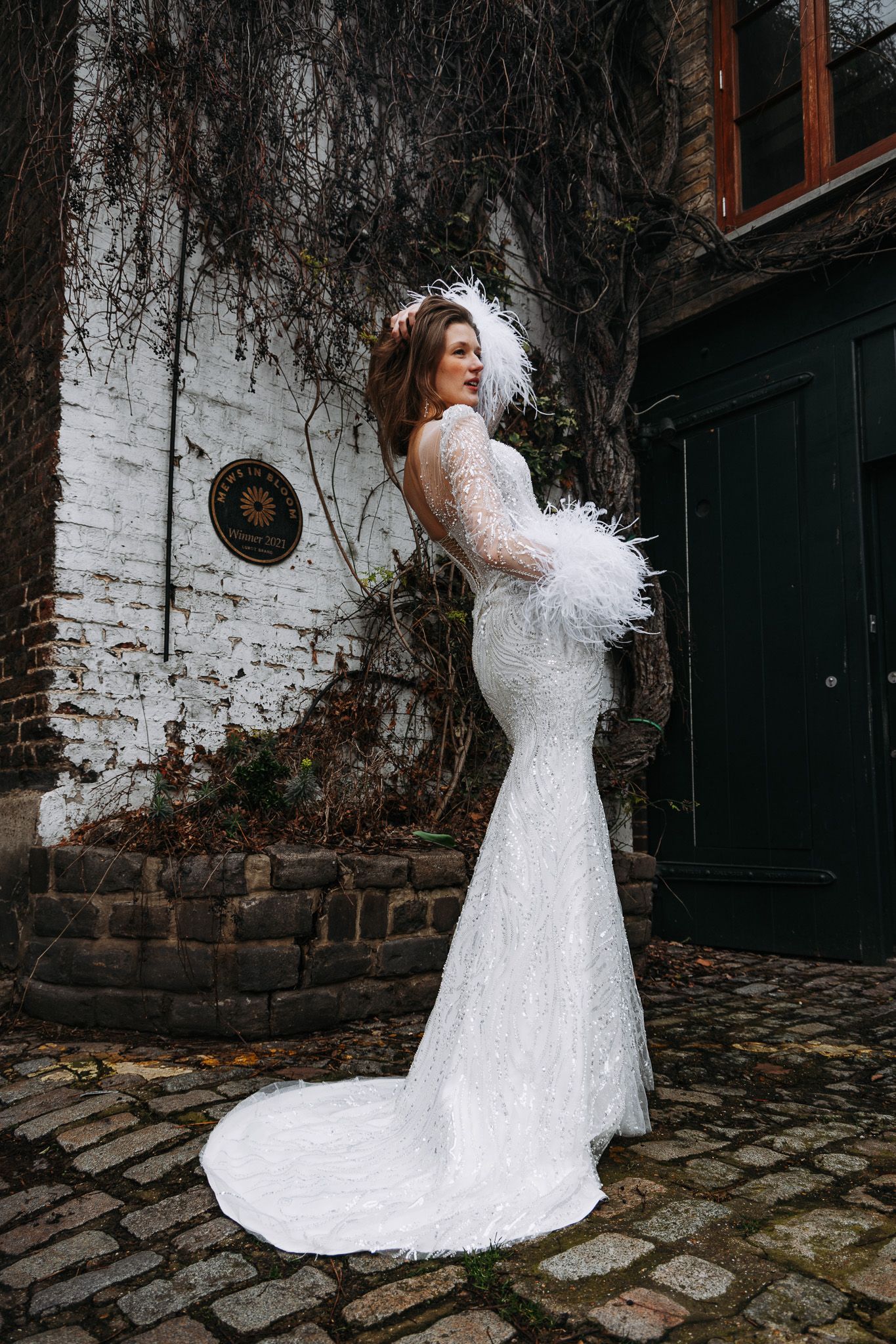A bride in a white lace gown with feather details poses against a rustic brick wall and cobblestone path.