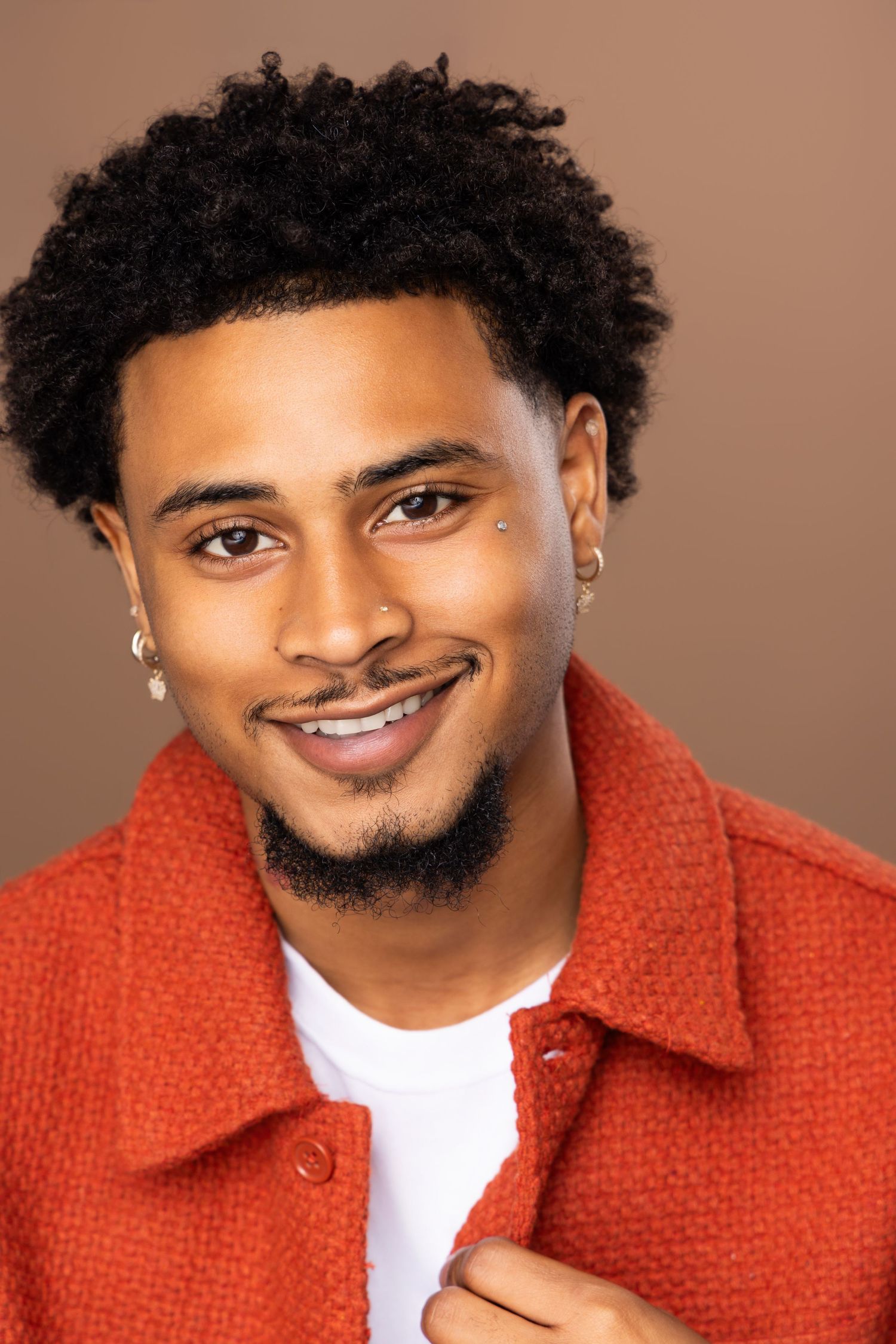 A person in a bright orange textured jacket and white t-shirt smiles warmly against a brown backdrop.