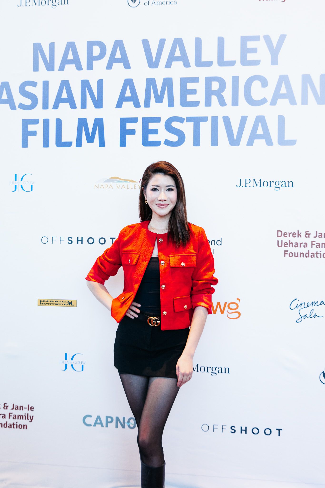 A person in a red jacket and black outfit poses at the Napa Valley Asian American Film Festival backdrop.