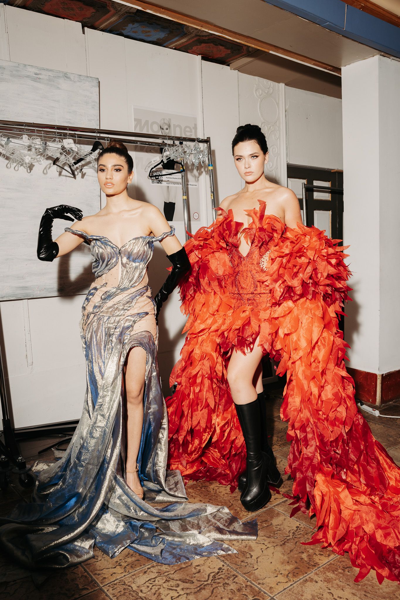 Fashion models pose in contrasting silver metallic and vibrant orange feathered gowns during an artistic photoshoot.