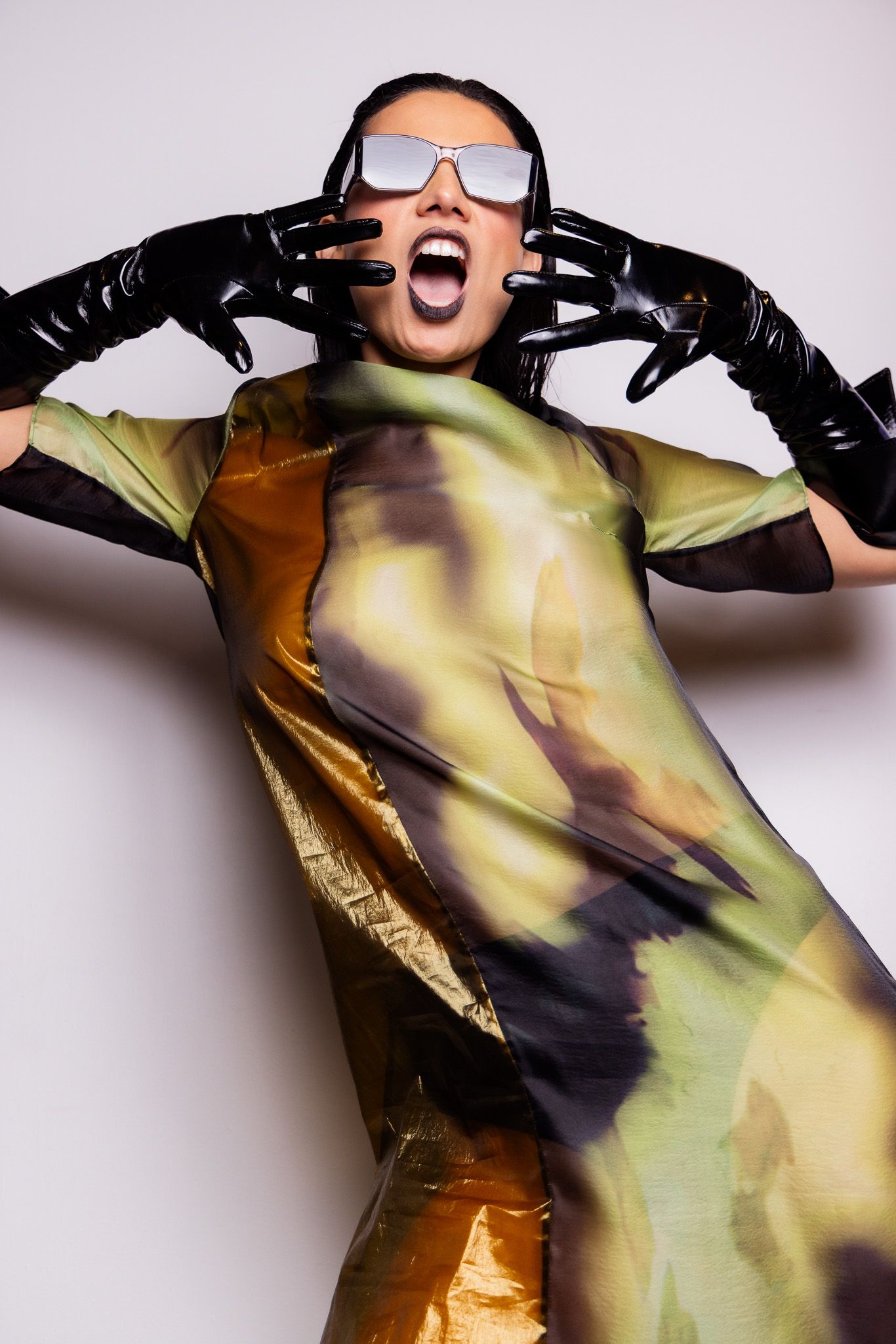 Model wearing iridescent yellow dress and black gloves poses expressively in futuristic sunglasses against white backdrop.